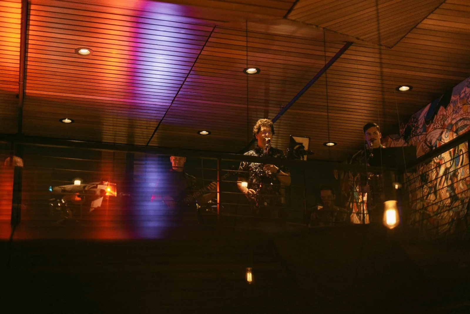 A band performs on an indoor balcony stage under warm, colorful lighting. Reflections from the lights create a vibrant atmosphere, and musicians are visible behind a railing. 