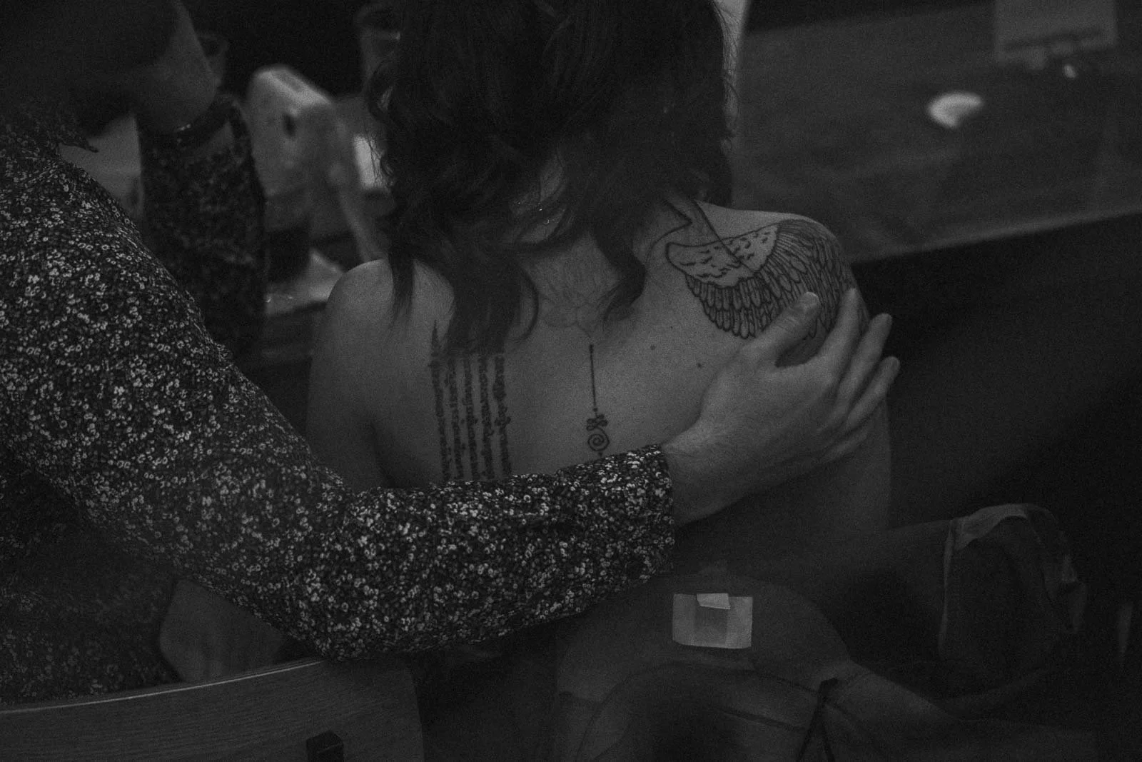  A person with a large wing tattoo on their shoulder and another tattoo on their back sits closely with another person, who has an arm around their back. The scene is in black and white and appears to be candid. 