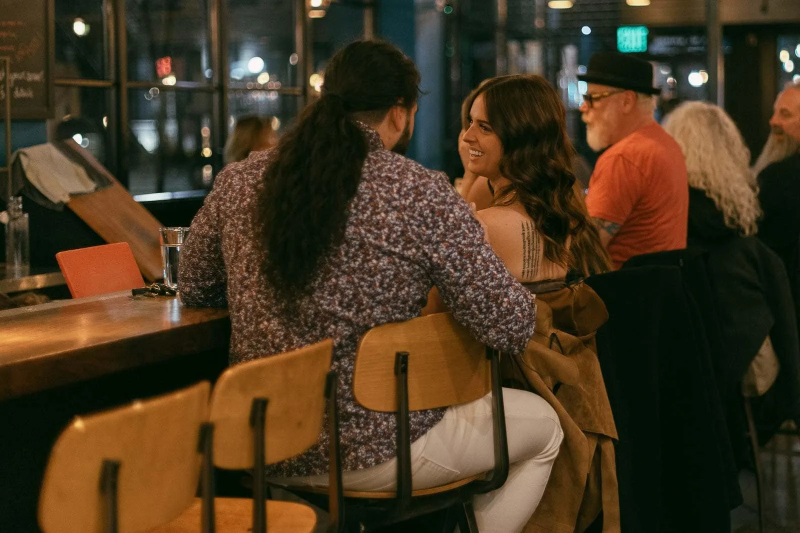  A man and woman sit close together at a bar, engaged in conversation and smiling. Other patrons are seated nearby in the dimly lit, cozy atmosphere. 