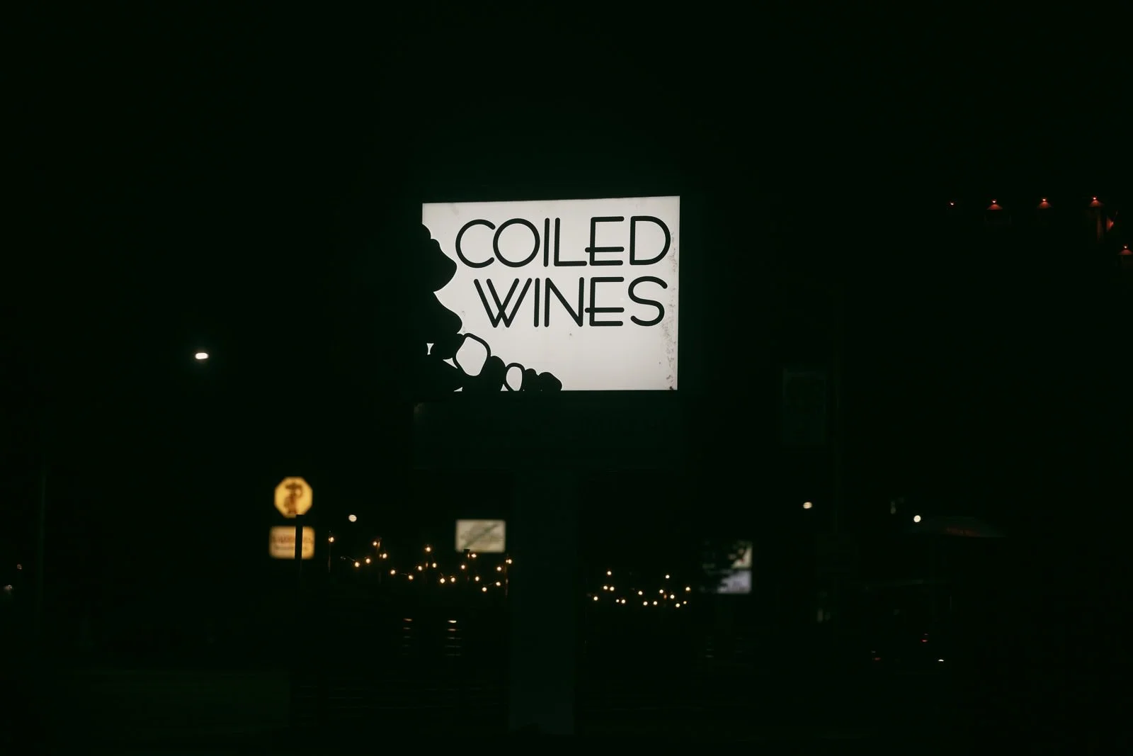  A brightly lit sign at night reads "COILED WINES" with a graphic of grape clusters on the left side, set against a dark background with faint lights visible in the distance. 