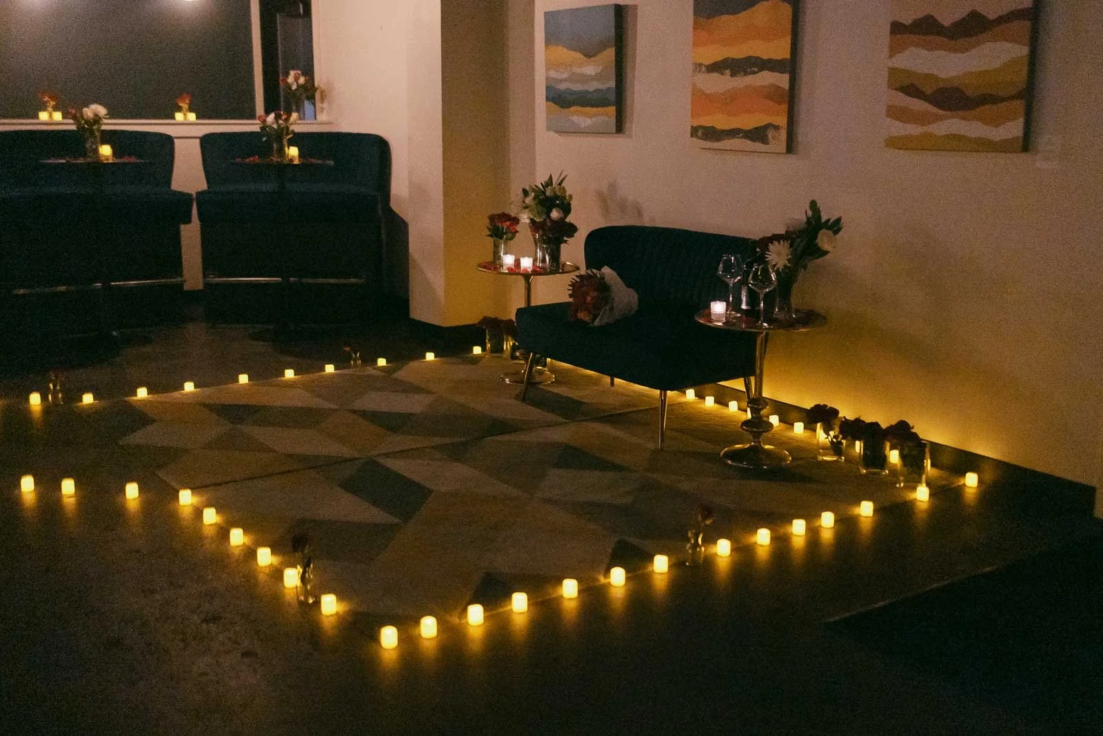  A cozy lounge area features a dark loveseat, flowers, wine glasses, and small tables surrounded by glowing LED candles on the floor. Abstract art hangs on the wall, creating a warm, intimate atmosphere. 