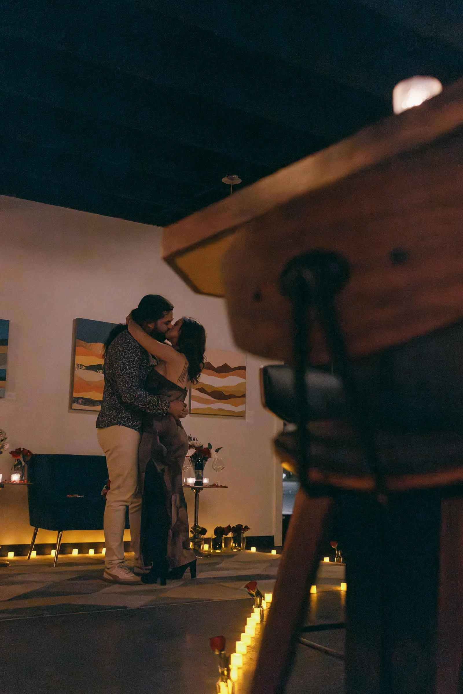  A couple embraces and kisses in a warmly lit room decorated with candles and rose petals, creating a romantic atmosphere. Artwork hangs on the wall behind them. 
