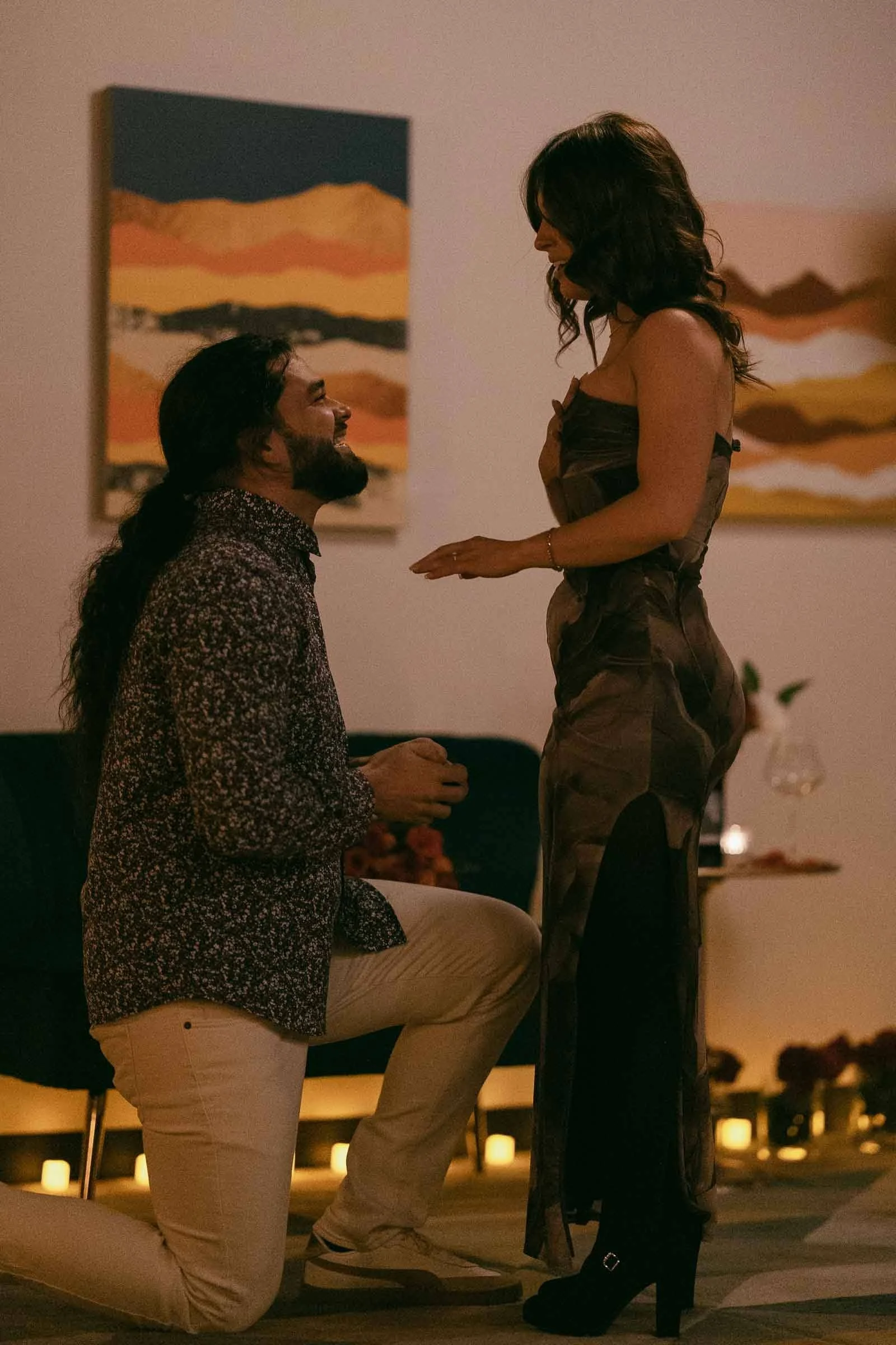  A man kneels in front of a woman, holding her hand, as if proposing. They are indoors, surrounded by candles, with abstract art on the wall behind them. Both appear happy and dressed for a special occasion. 