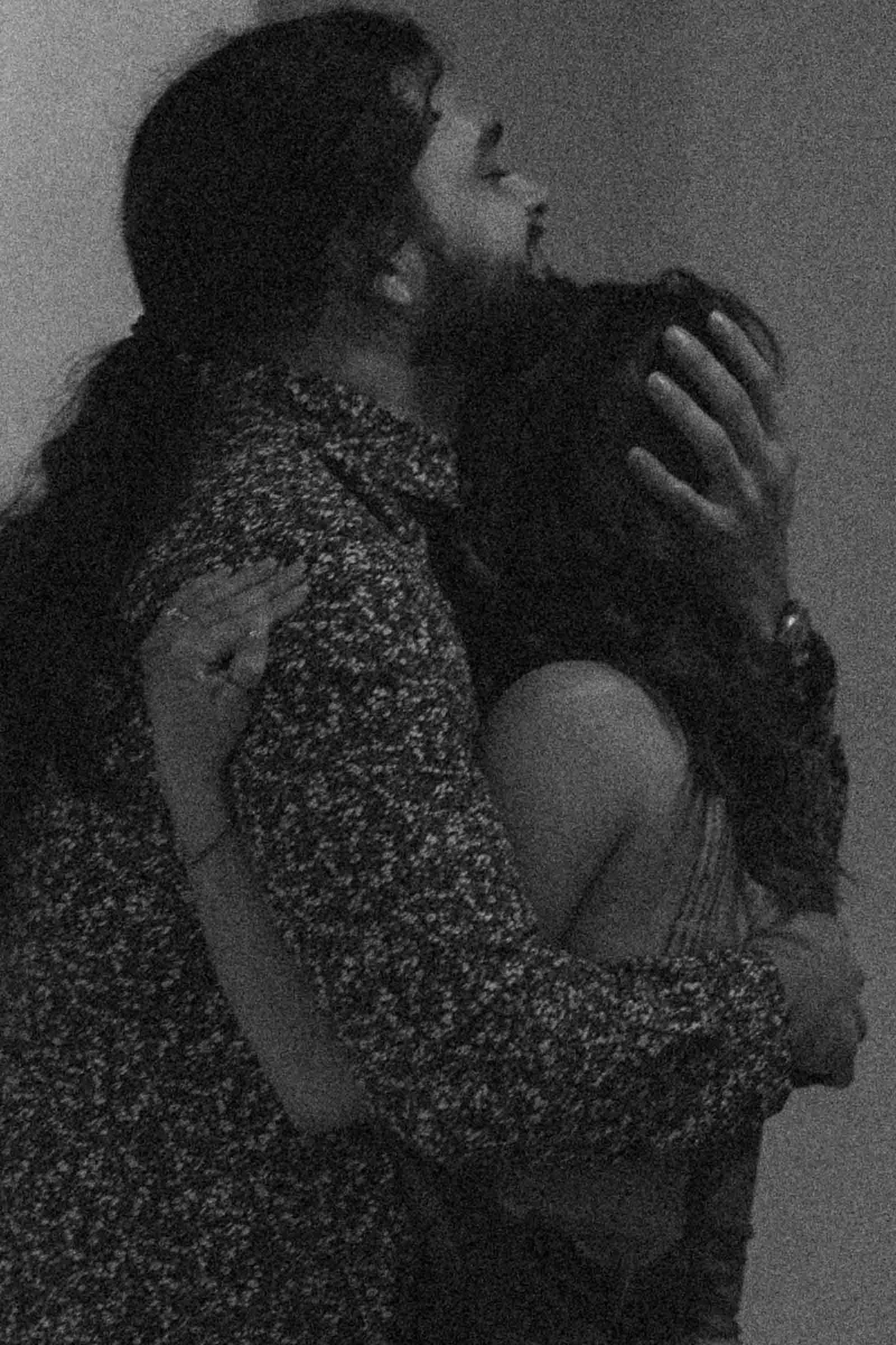  A man with long hair, wearing a patterned shirt, embraces a woman, gently holding her head. The image is in black and white with a grainy texture, evoking a sense of intimacy and emotion. 