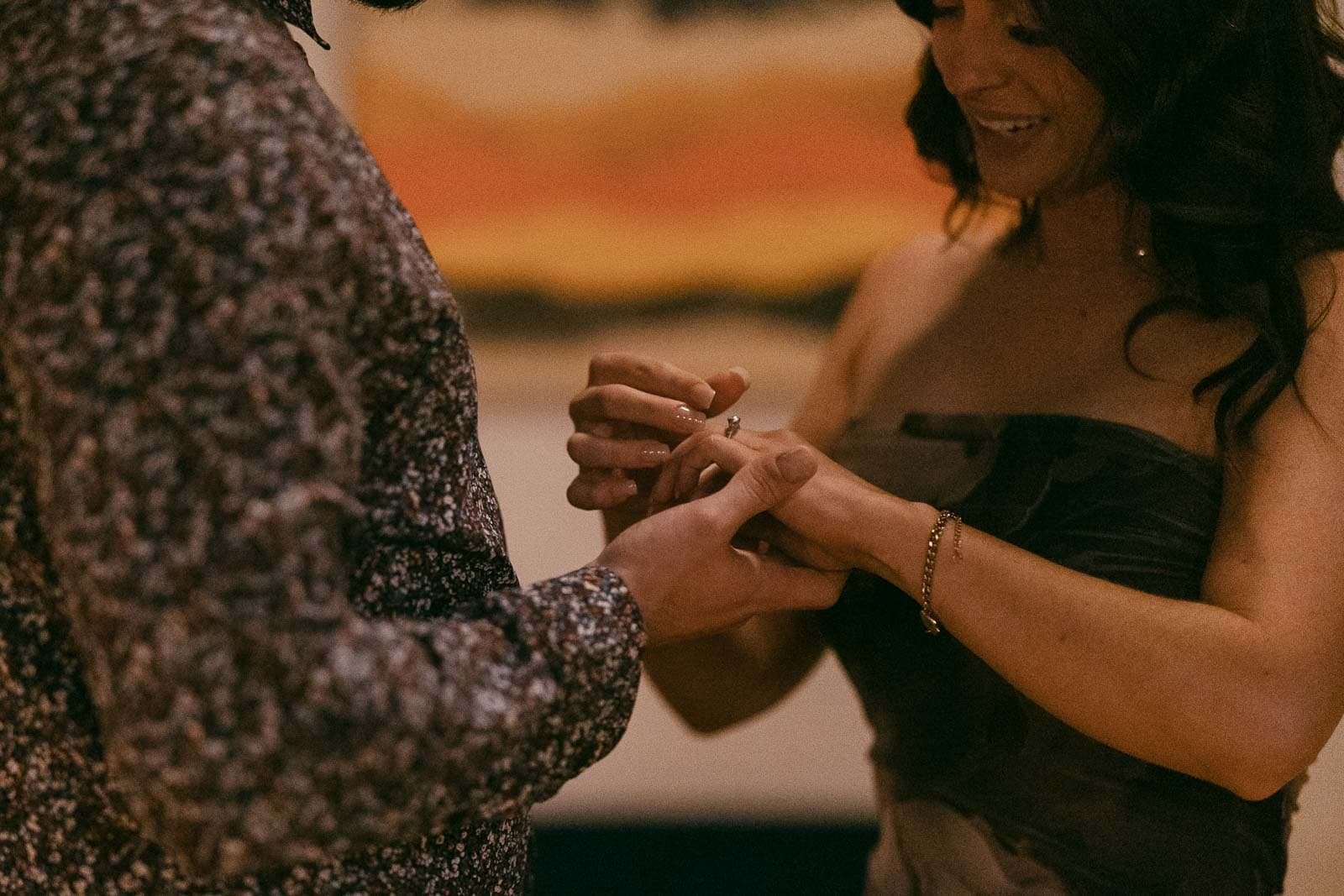  A woman in a strapless dress places a ring on the finger of a person in a patterned shirt, both smiling and holding hands in a warm, intimate moment. 