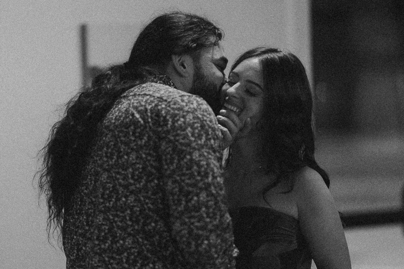 A man with long hair and a patterned shirt leans in to kiss a smiling woman with long dark hair and a strapless dress. He touches her face affectionately. The black-and-white photo has a candid, intimate feel.