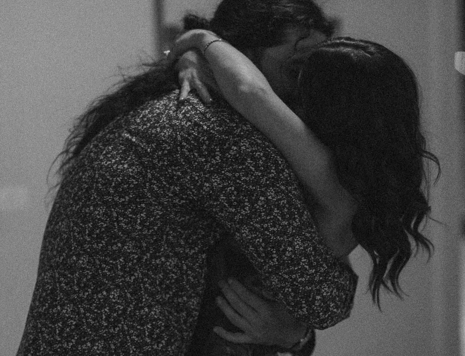  Two people embrace closely in a black and white photo. One person has long, wavy hair and both have their arms wrapped tightly around each other, conveying a sense of intimacy and affection. 