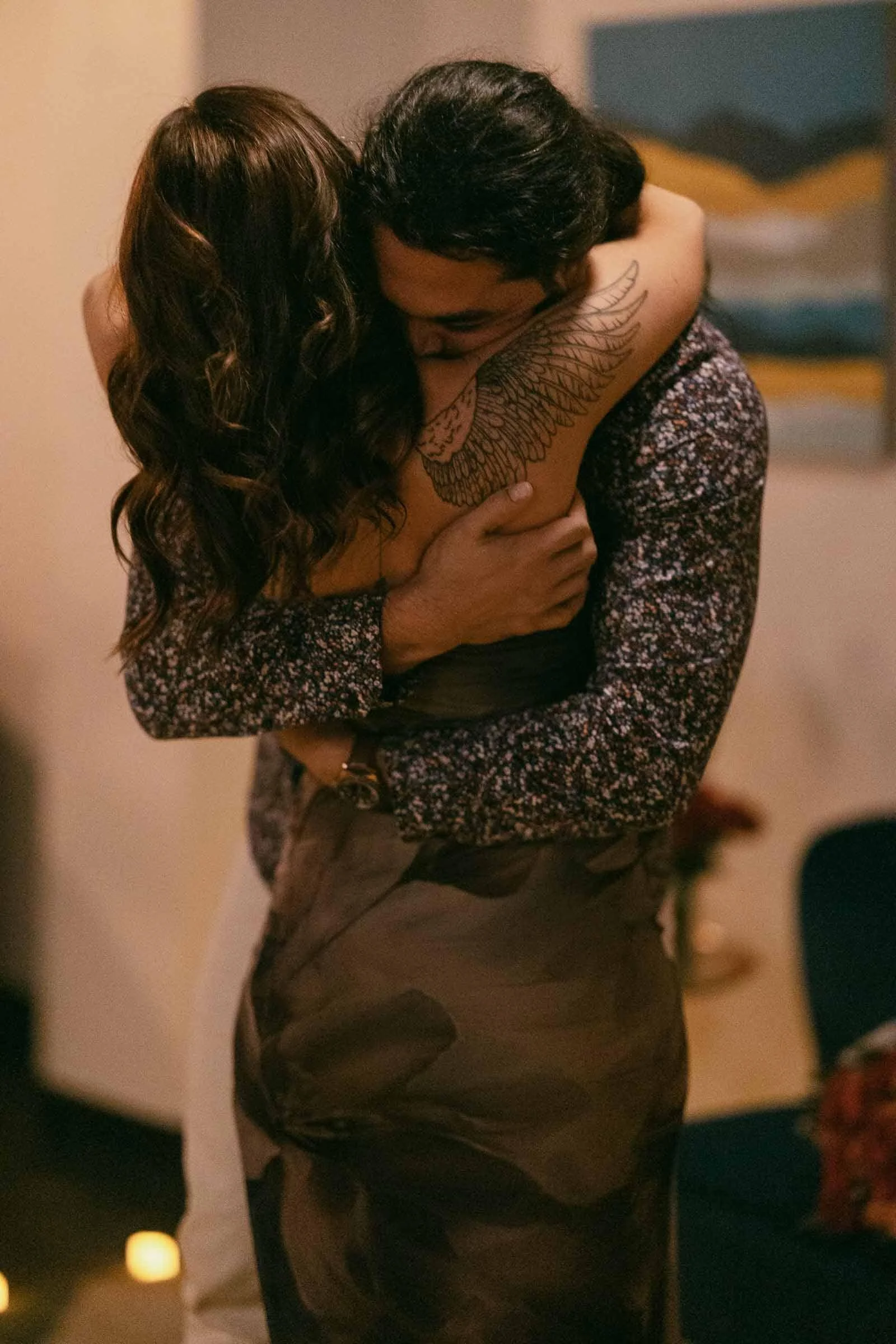  A couple embraces closely in a warmly lit room. The woman, with wavy hair and a wing tattoo on her arm, wears a sleeveless dress while the man, in a patterned shirt, hugs her tightly. A blurred painting hangs in the background. 