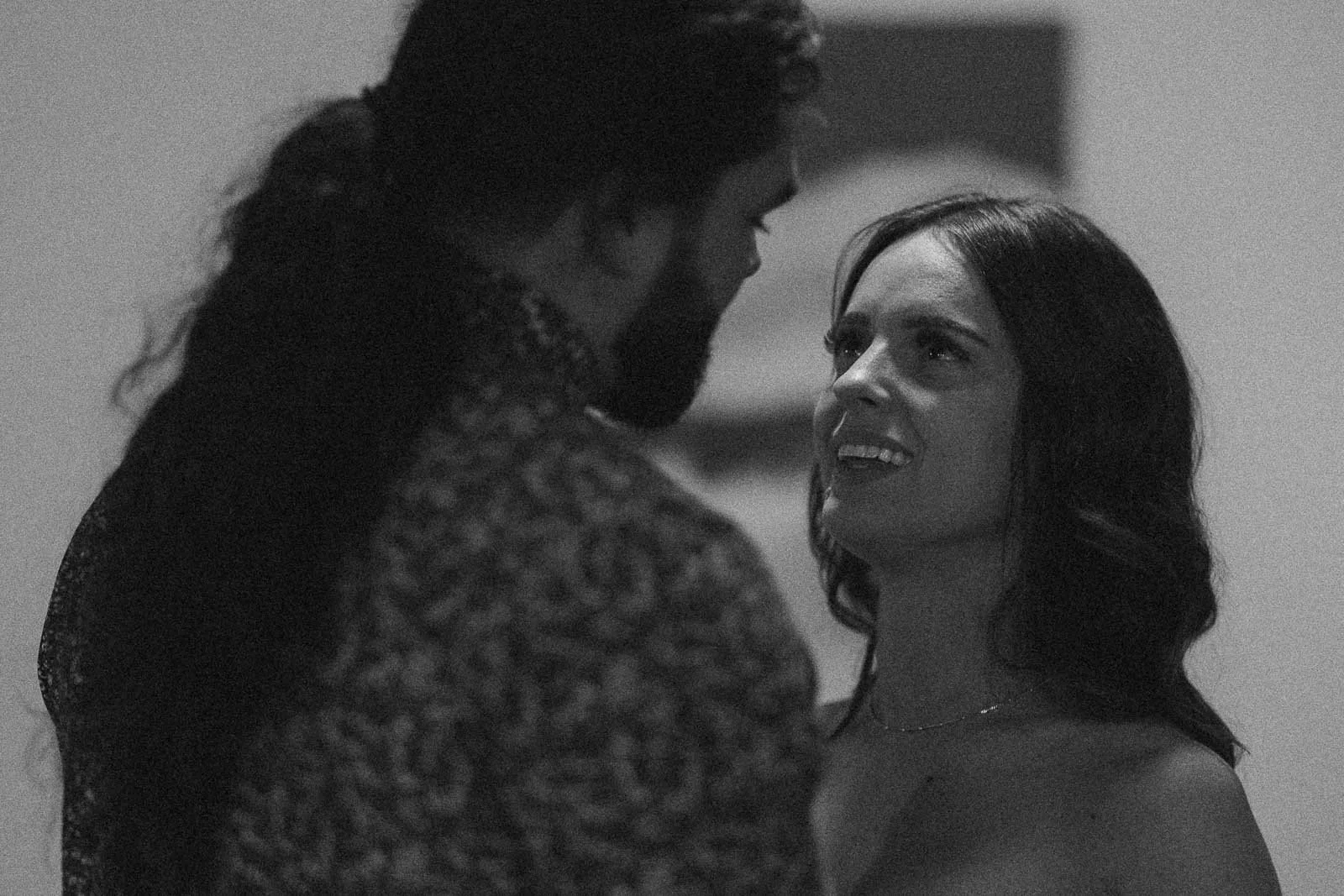  A black-and-white photo of a woman smiling and looking up at a man with long hair. They are standing close together indoors, appearing to share an intimate moment. 