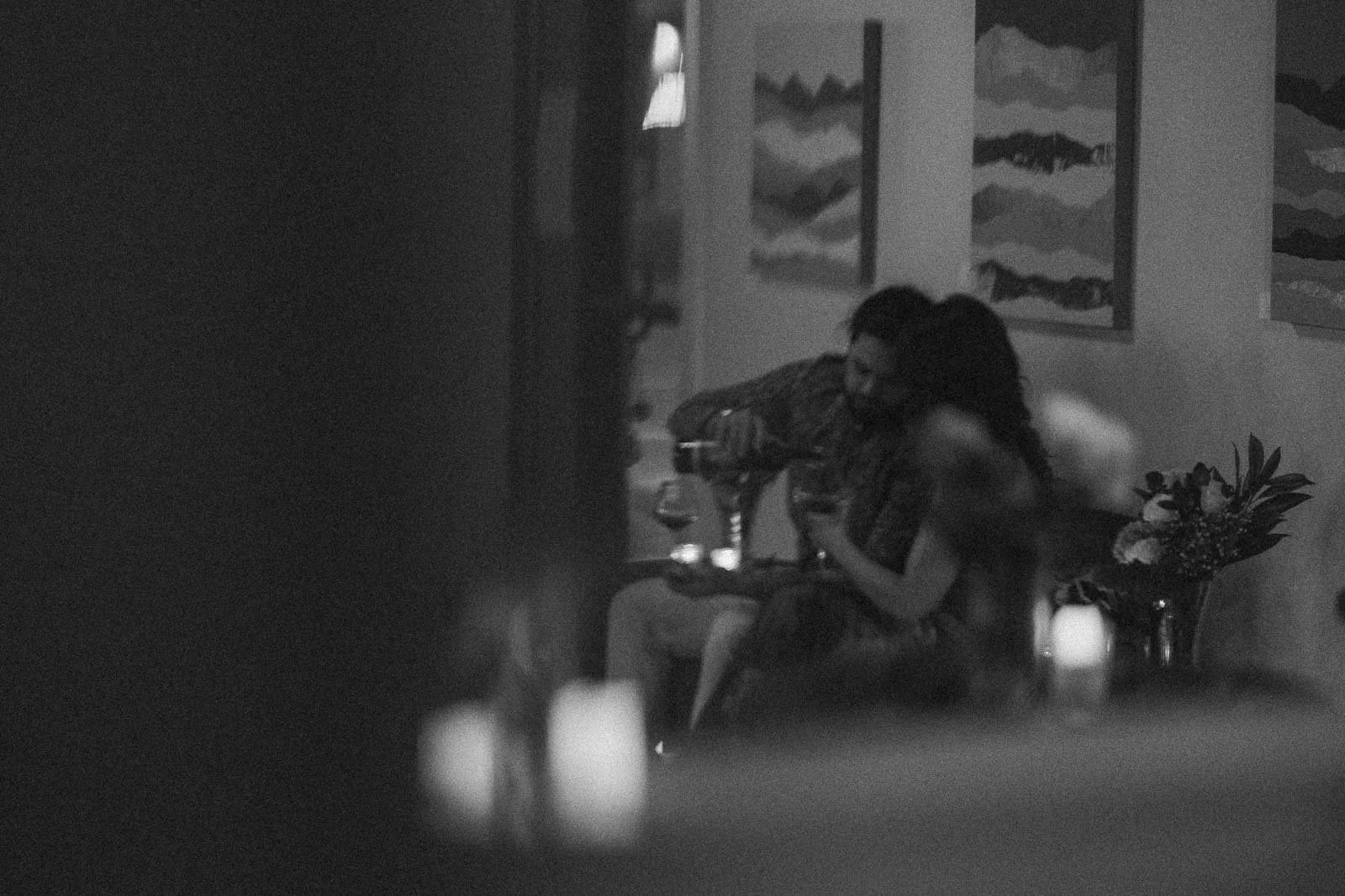  A black-and-white, softly blurred photo of a couple sitting closely together indoors, with one person pouring wine into a glass. There are abstract paintings and a bouquet of flowers in the background. 