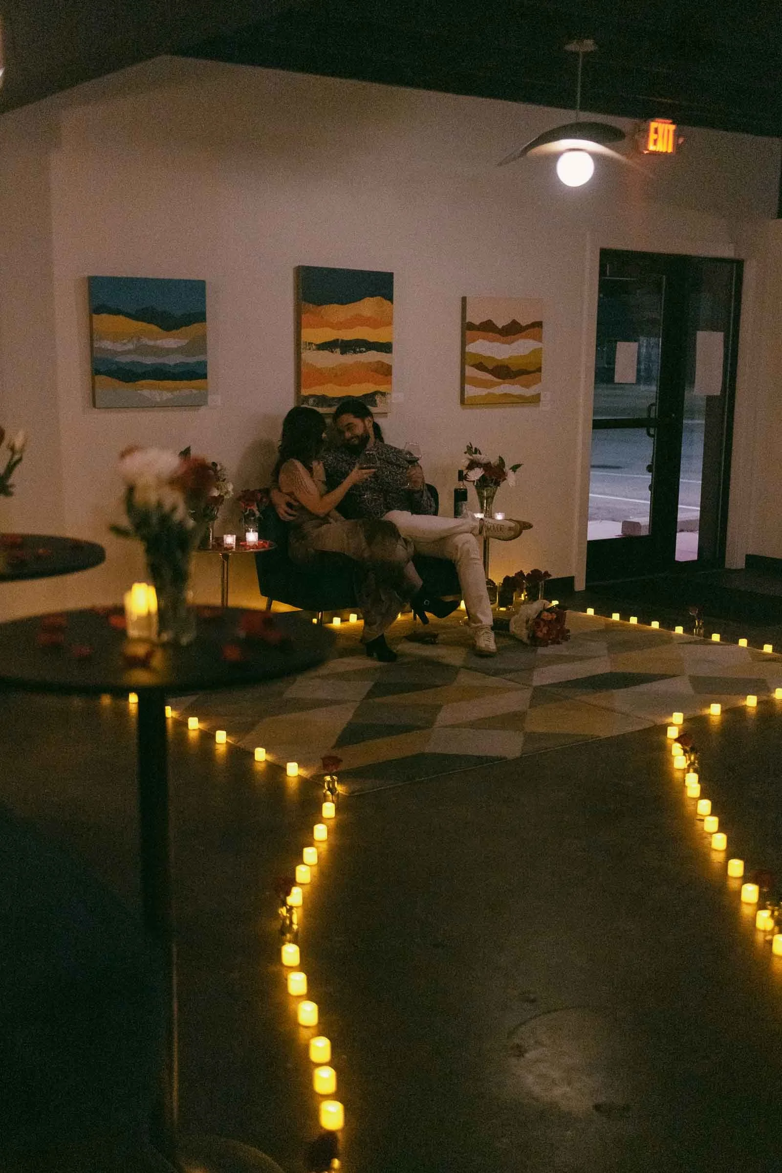  A couple sits closely together on a bench in a dimly lit room decorated with candles forming a heart shape on the floor, flowers on tables, and landscape paintings on the wall, creating a romantic atmosphere. 