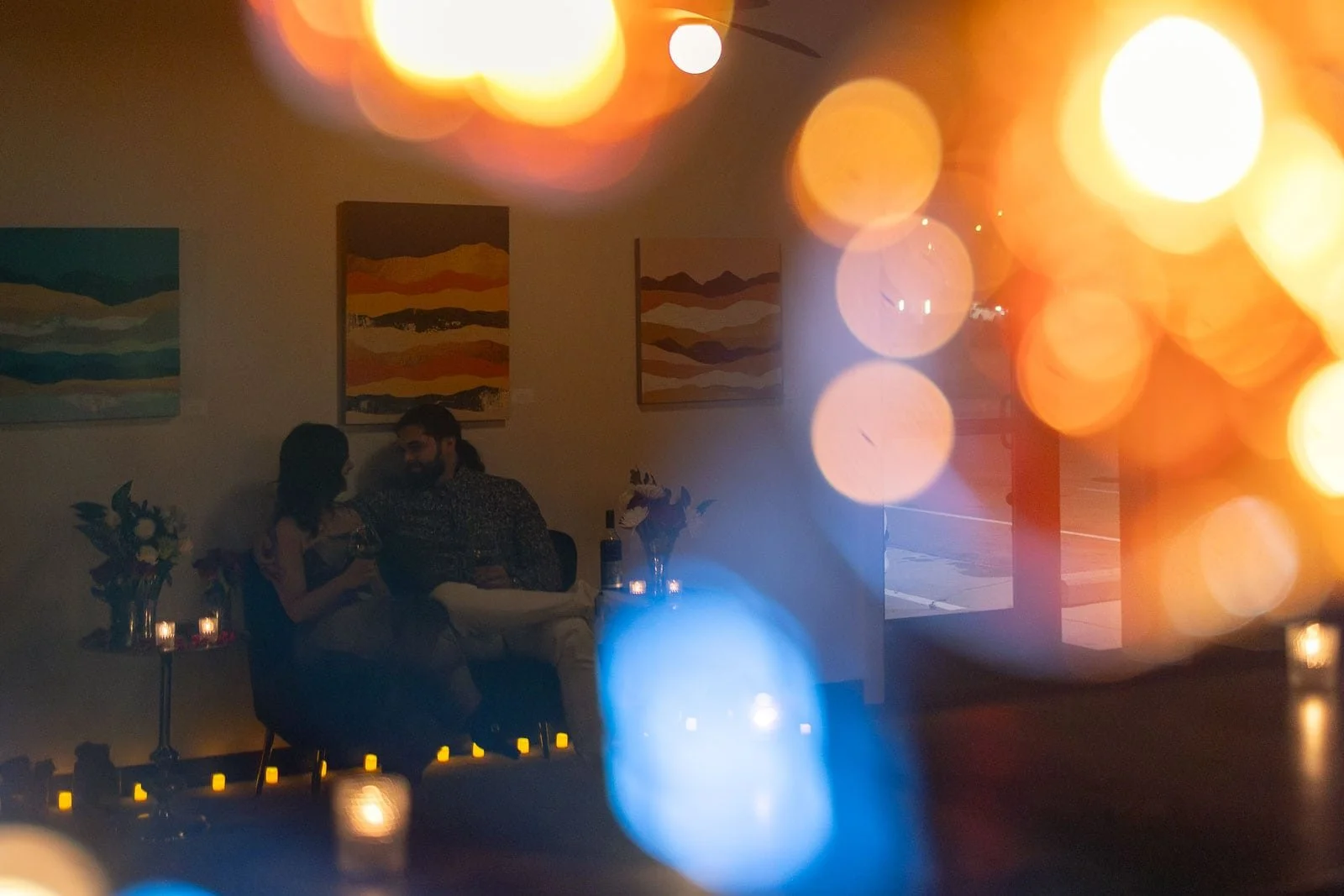 A couple sits closely together in at Coiled Wines, surrounded by candles and abstract paintings. The image is double exposed with warm, soft bokeh lights from Funky Taco creating a dreamy, romantic atmosphere.