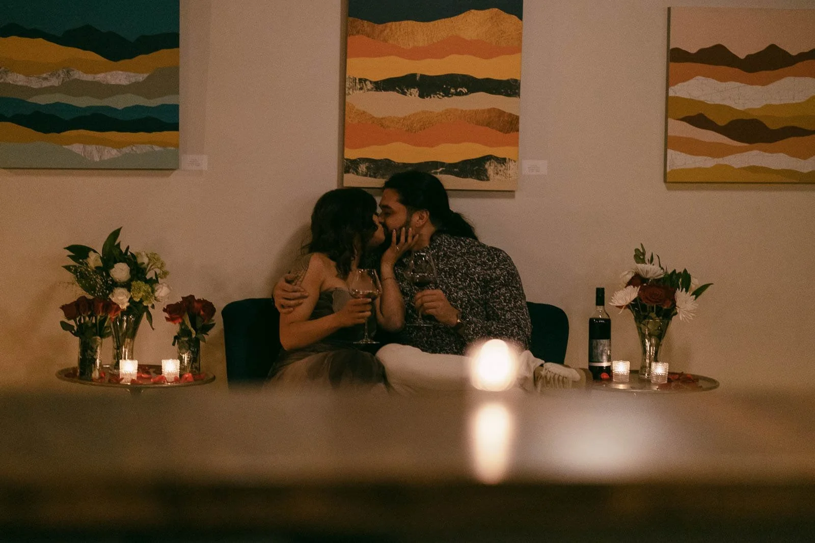  A couple sits on a couch, sharing a kiss while holding glasses of wine. They are surrounded by flowers, candles, and wine bottles, with colorful abstract art hanging on the wall behind them. The man has just proposed to his new fiancée at the locati
