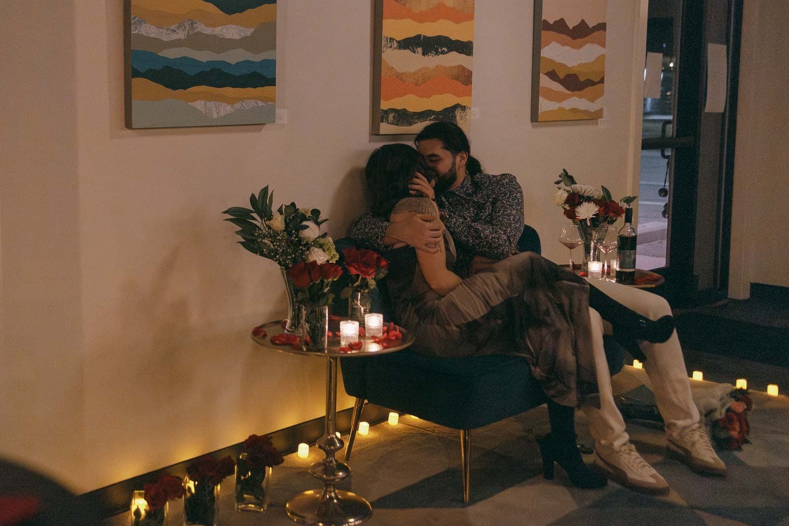  A couple sits closely together in a dimly lit room, surrounded by candles and flowers. They embrace and kiss on a blue chair near a small table with wine glasses and lit candles, creating a romantic atmosphere. 