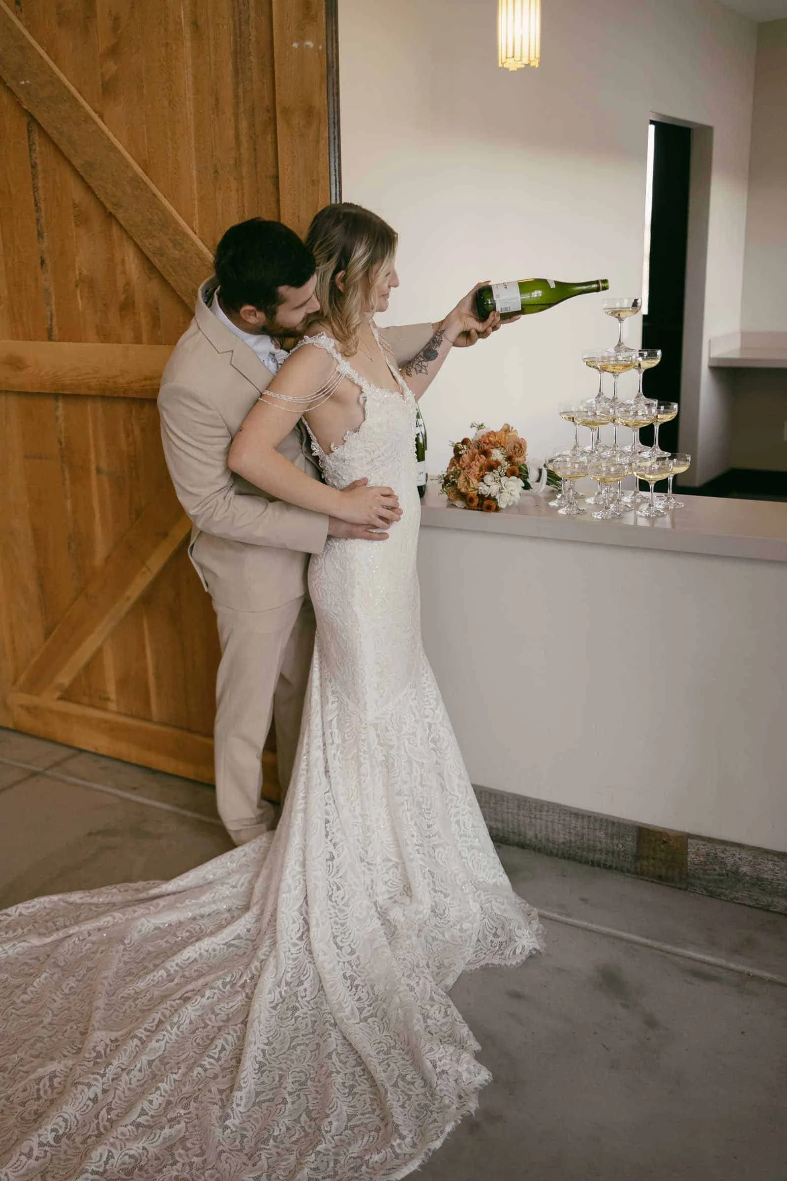  A bride in a lace gown pours champagne into a tower of glasses, while the groom in a beige suit stands behind her, embracing her waist. They are indoors near a wooden door and a bouquet of flowers sits by the glasses. 