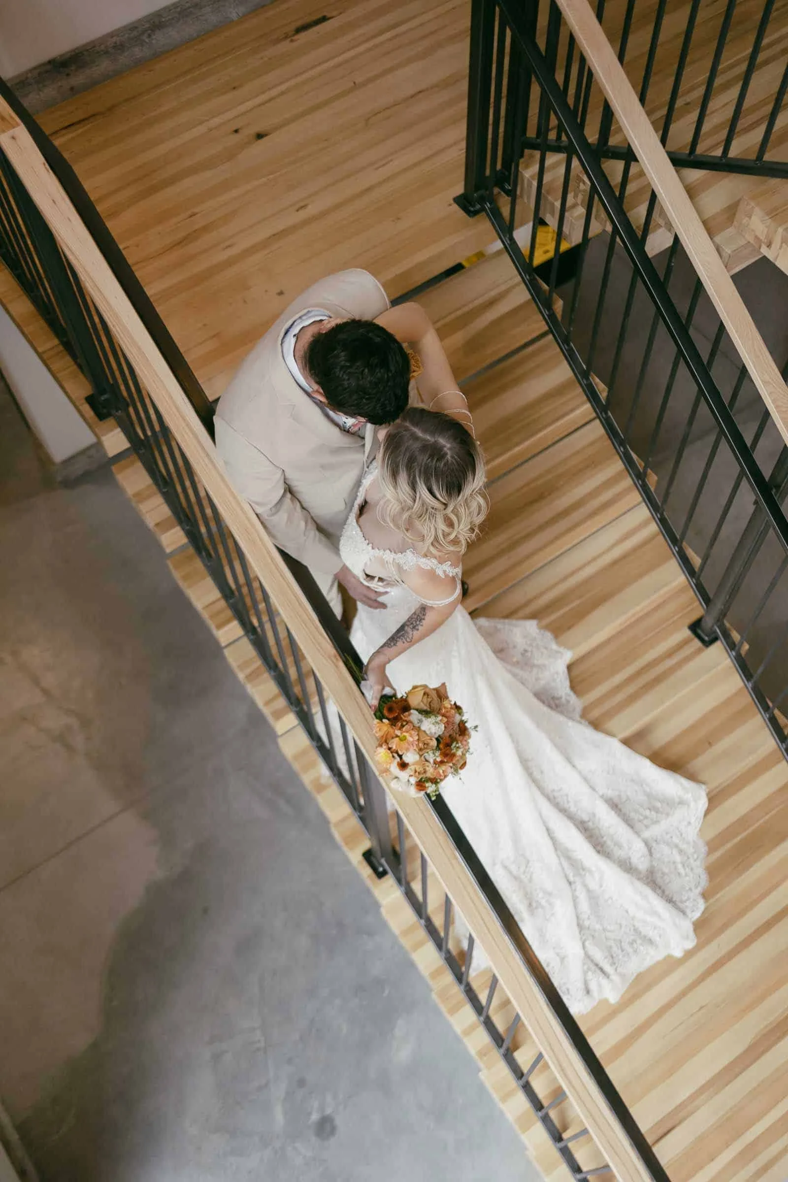  A bride in a white gown and a groom in a light suit embrace on a wooden staircase, viewed from above. The bride holds a bouquet of flowers and both are looking lovingly at each other. 