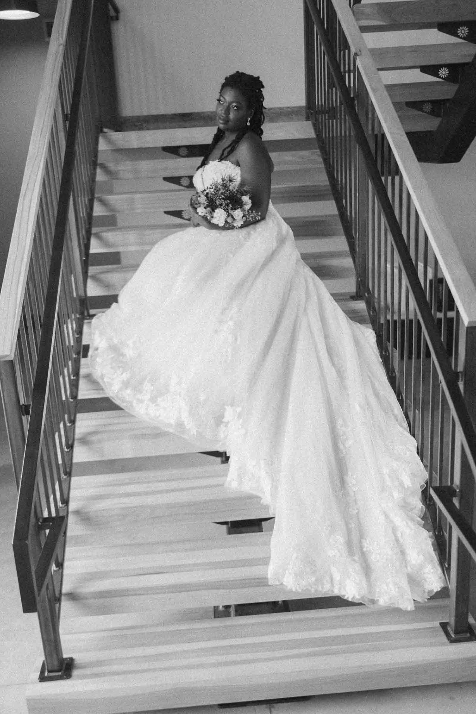  A bride lies across a wooden staircase adorned in a sparkly wedding dress. She looks out the window, preparing for her wedding day. The image is in black and white 