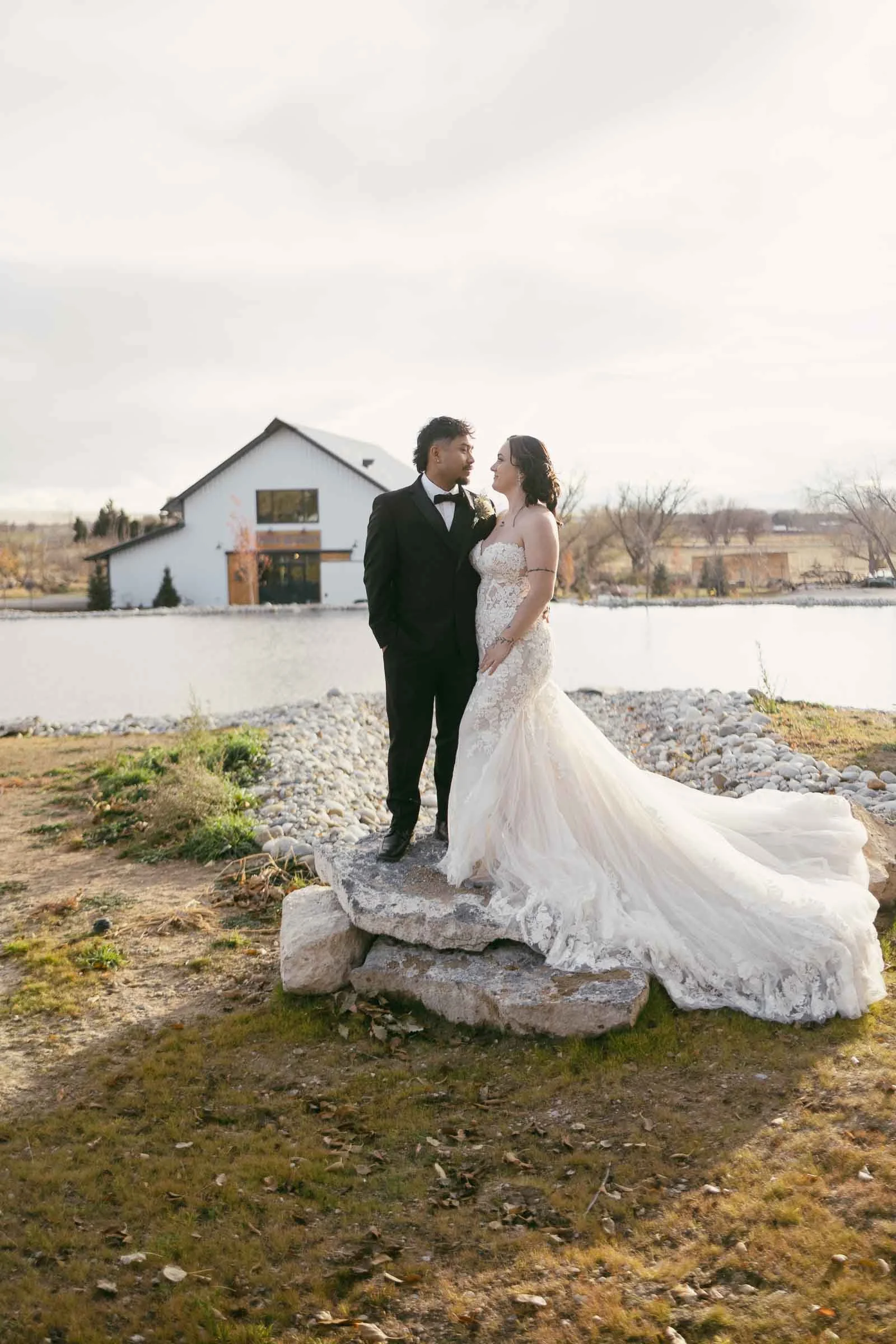  A bride in a lace wedding gown and a groom in a black tuxedo stand together on rocks near a pond, with a white barn-style building in the background under a bright sky. 