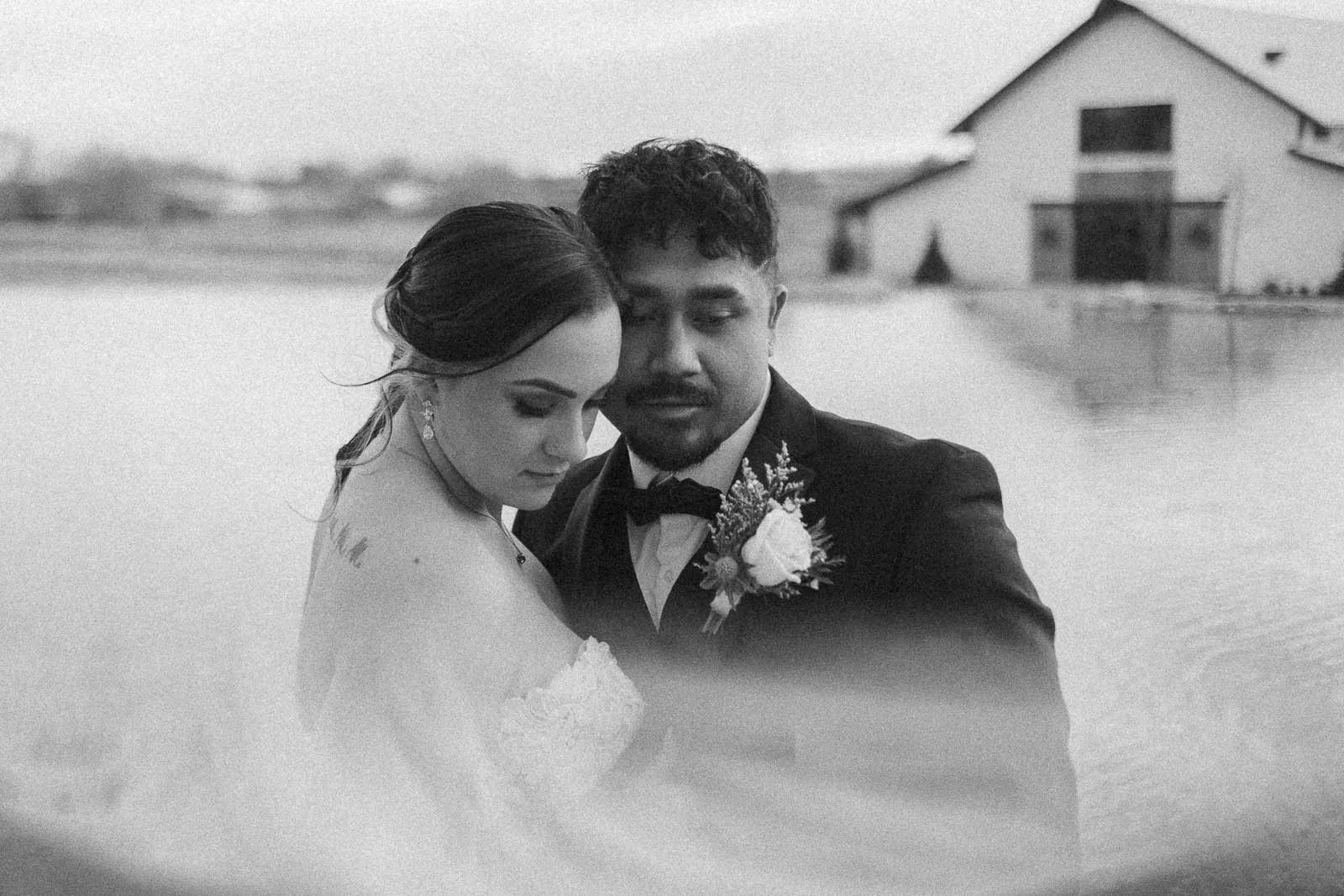  A bride and groom embrace closely by the water, with a large barn and blurred landscape in the background. The image is black and white, creating a soft and romantic atmosphere. 