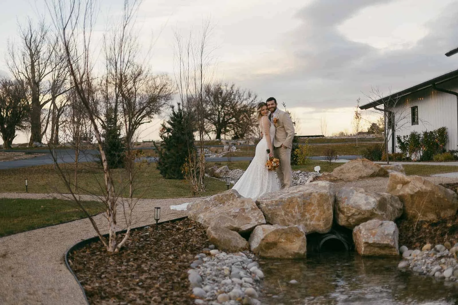  A bride and groom in wedding attire embrace and smile beside large rocks and a small pond in a landscaped outdoor setting, with trees and a white building in the background. 