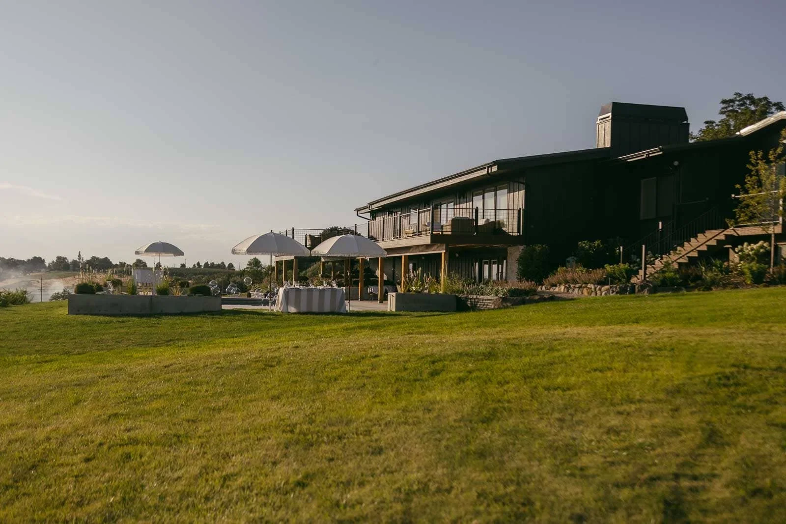  A modern building with large windows overlooks a spacious grassy lawn, with white umbrellas and outdoor seating arranged nearby under a clear sky. 