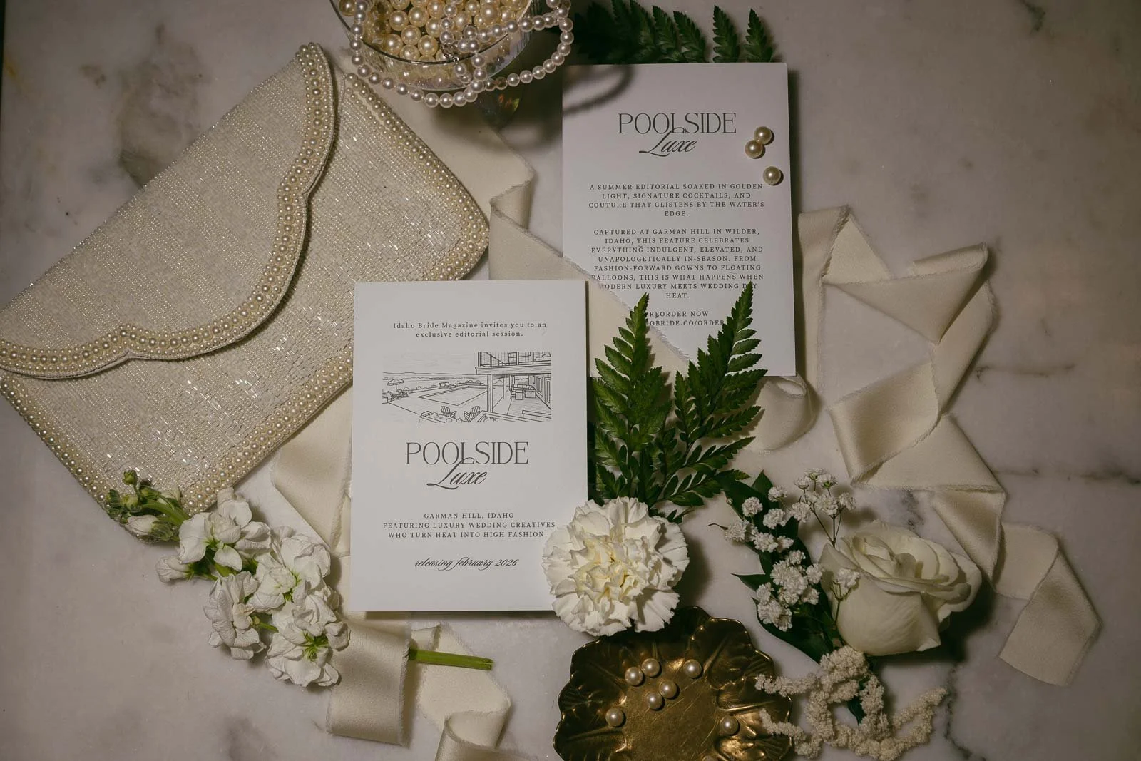  Wedding invitations with "Poolside Luxe" text, surrounded by white flowers, green fern leaves, a pearl-embellished clutch, a gold tray with pearls, and cream ribbons on a marble surface. 