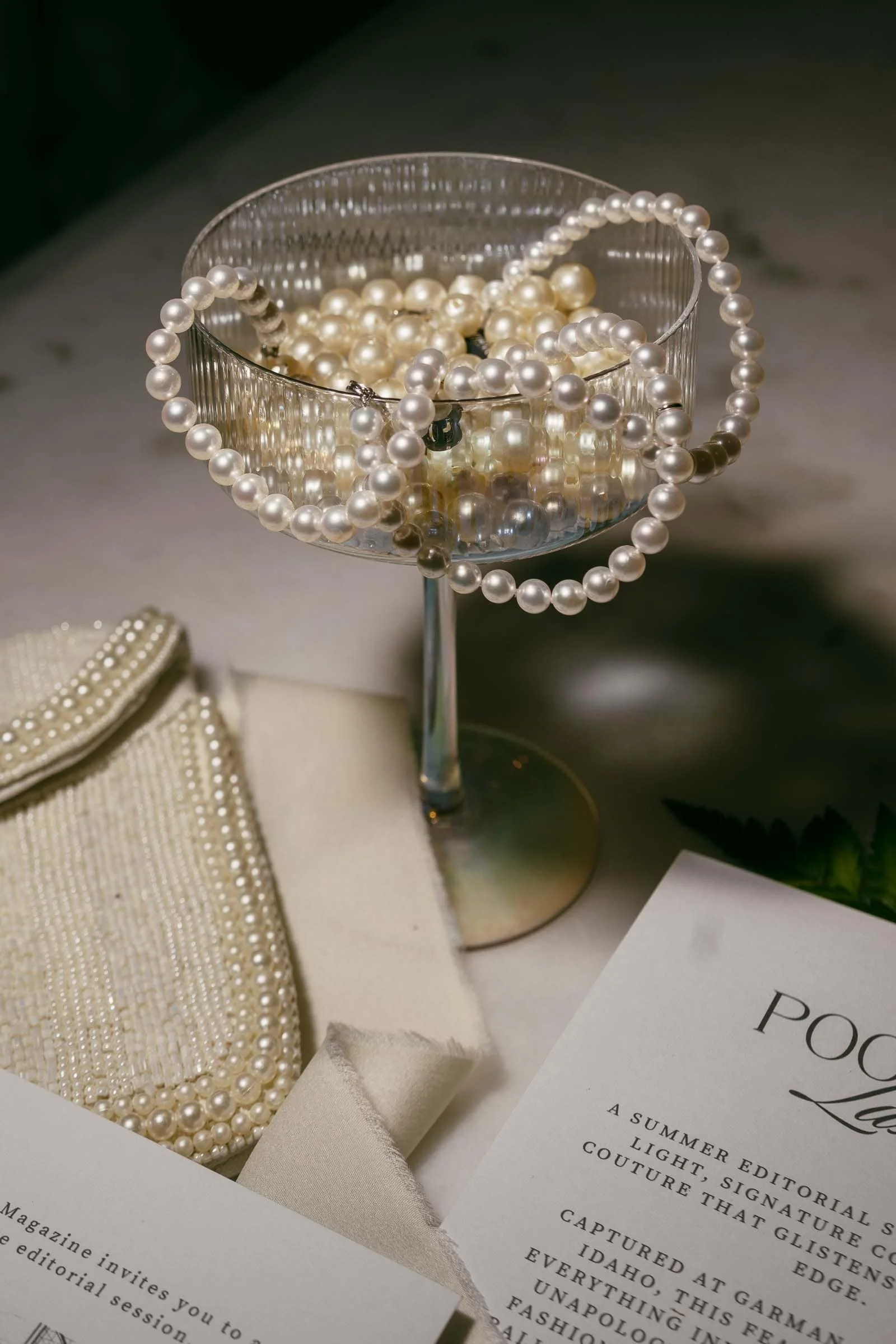  A glass coupe holds strands of white pearls, with more pearls draped over the rim. Nearby are a beaded clutch, cream fabric, and pages from a fashion magazine on a marble surface. 