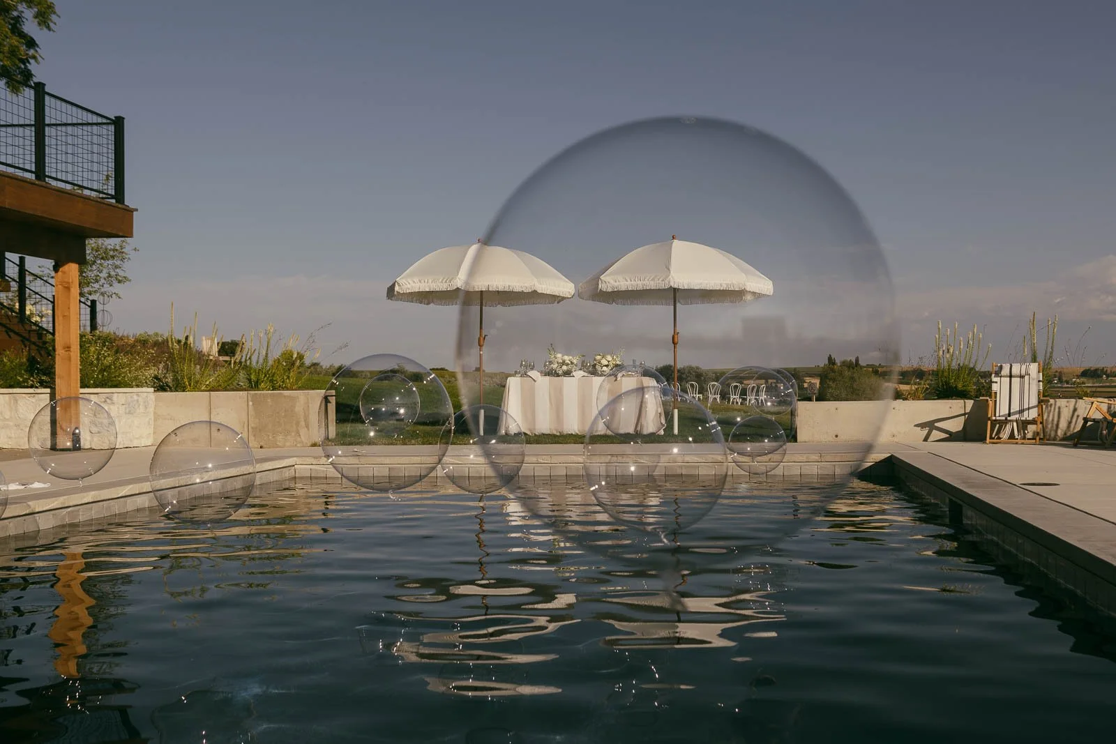  Large transparent bubbles float above a calm outdoor pool, with two white umbrellas and a table decorated for an event in the background under a clear sky. 