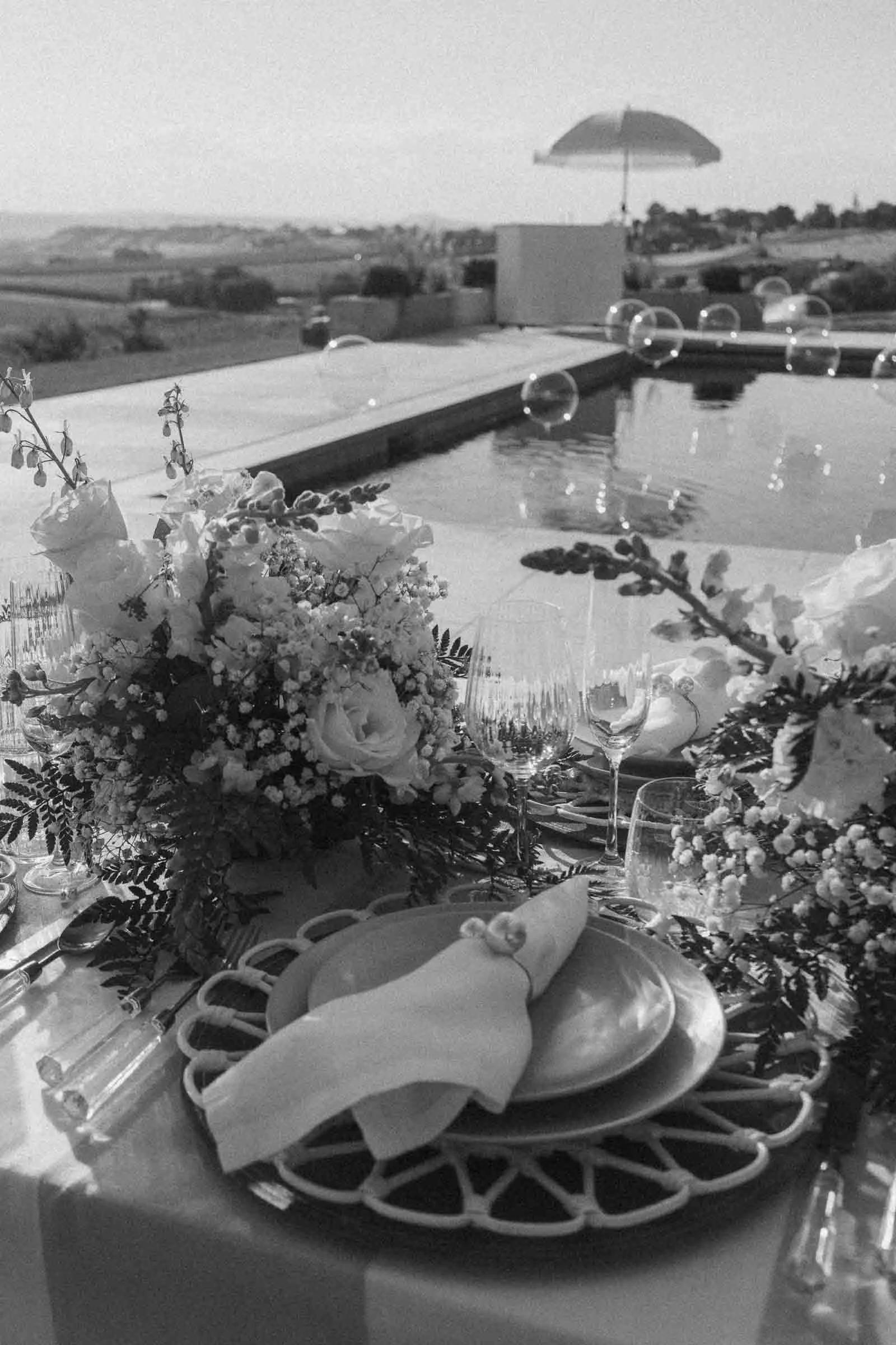 A beautifully set outdoor dining table with elegant plates, napkins, floral arrangements, and glasses beside a pool. In the background, bubbles float in the air and a landscape stretches under an open umbrella. 