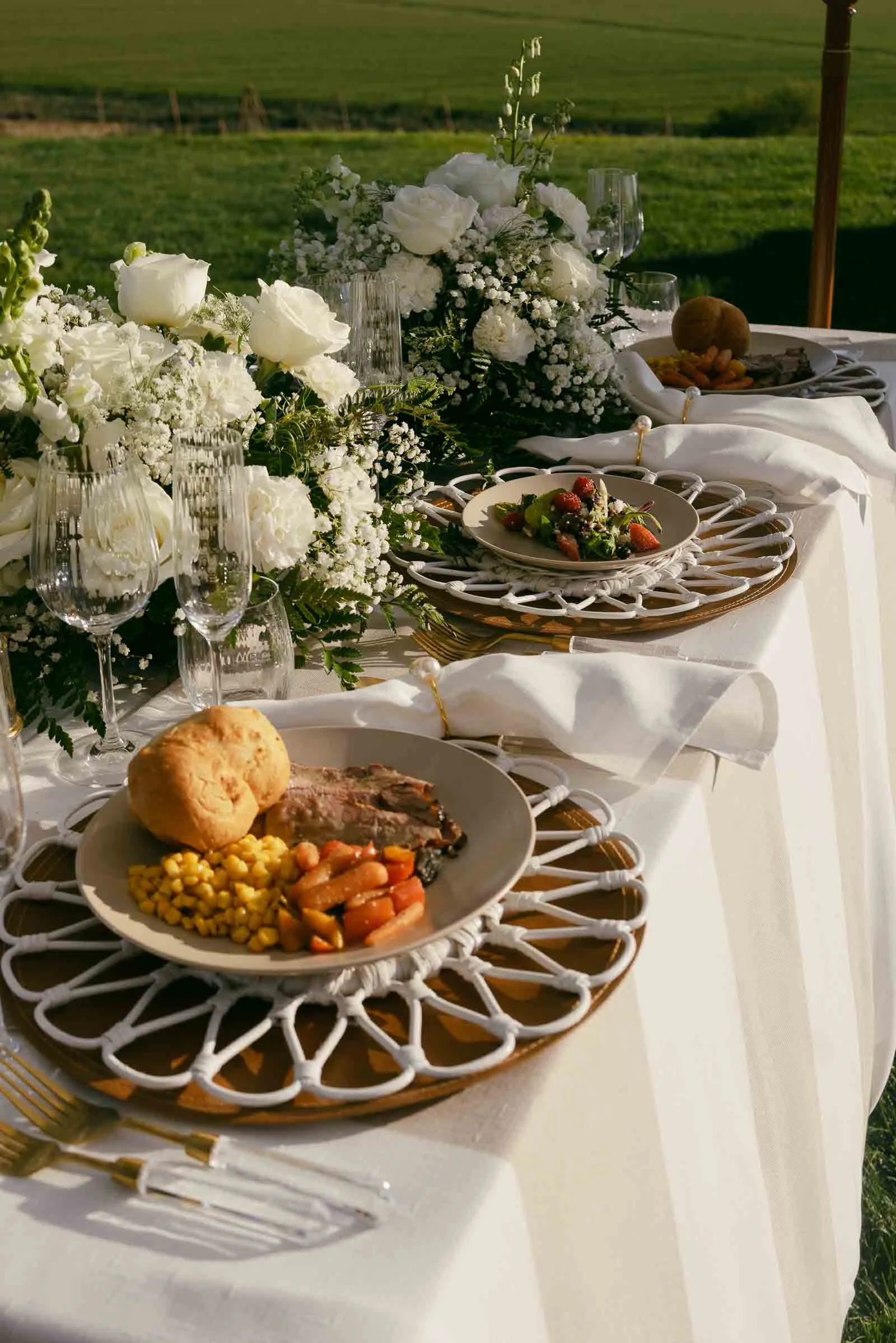  A beautifully set outdoor table features plates of food with roast beef, carrots, corn, and a roll, surrounded by elegant white floral arrangements and glassware, with a green field in the background. 