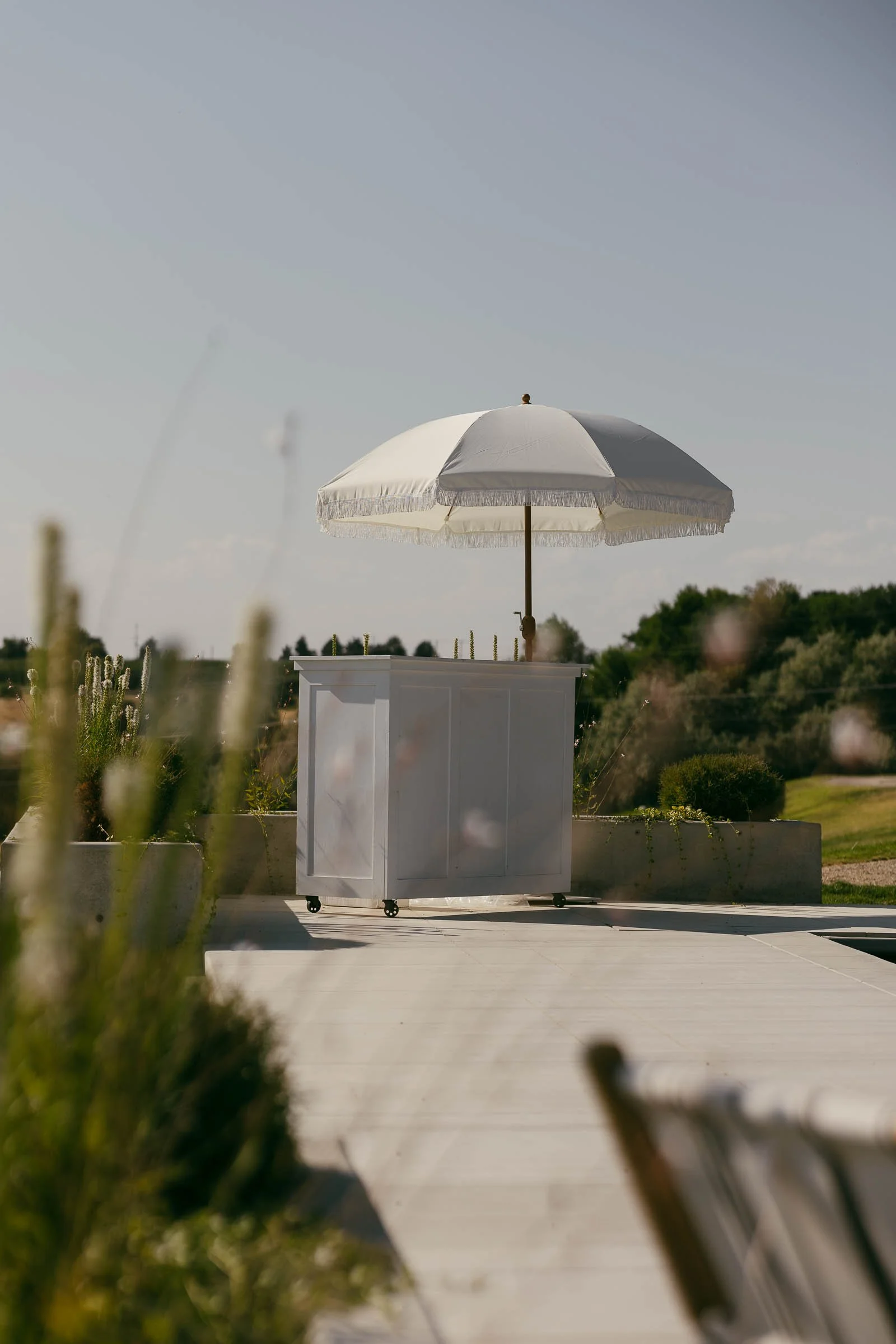  A small white bar cart with a white parasol stands outdoors on a sunny day, surrounded by greenery and a clear sky, with blurred plants in the foreground. 