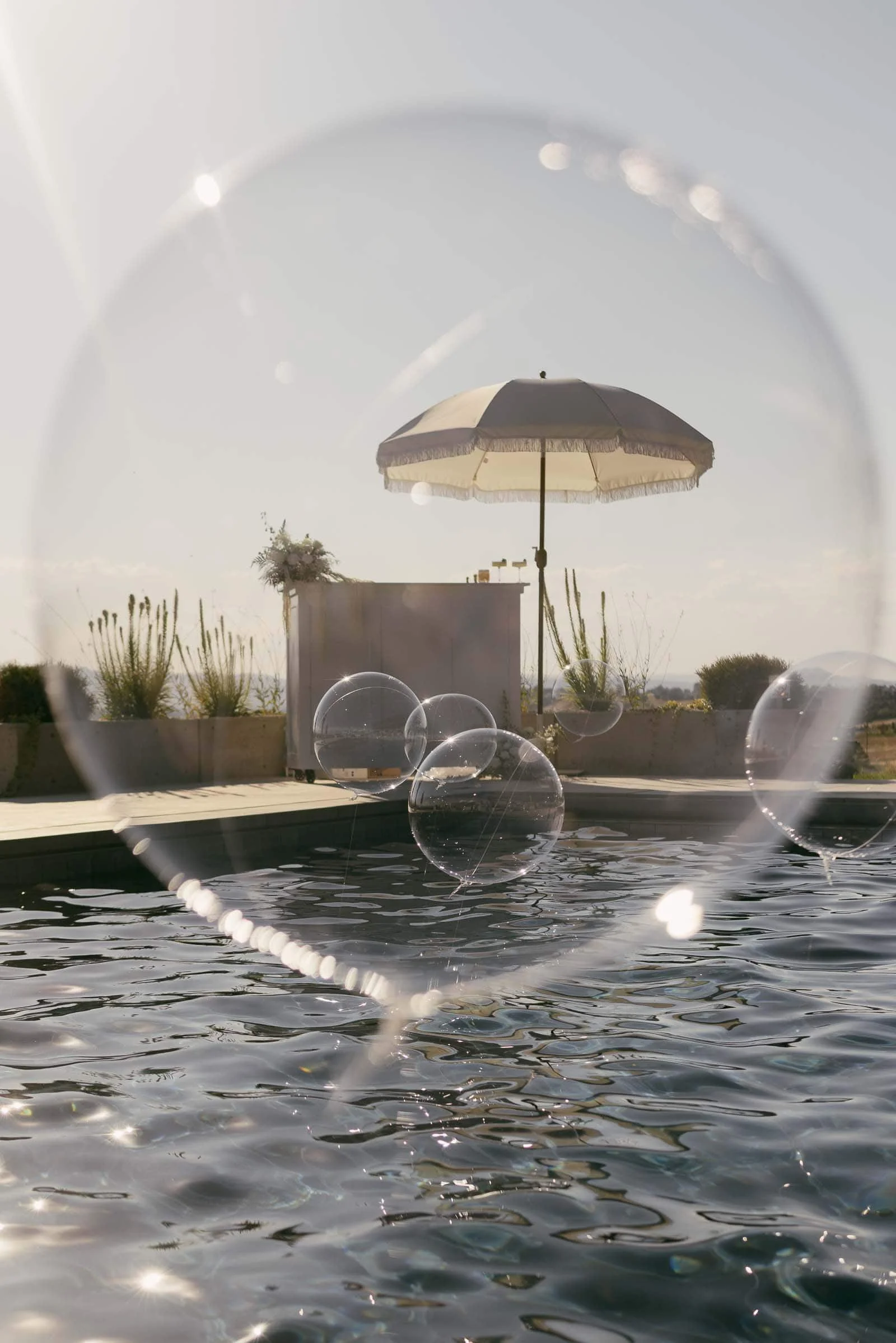  Large soap bubbles float above a rippling pool, with a white parasol shading a small bar or table in the background, all under soft sunlight on a clear day. 