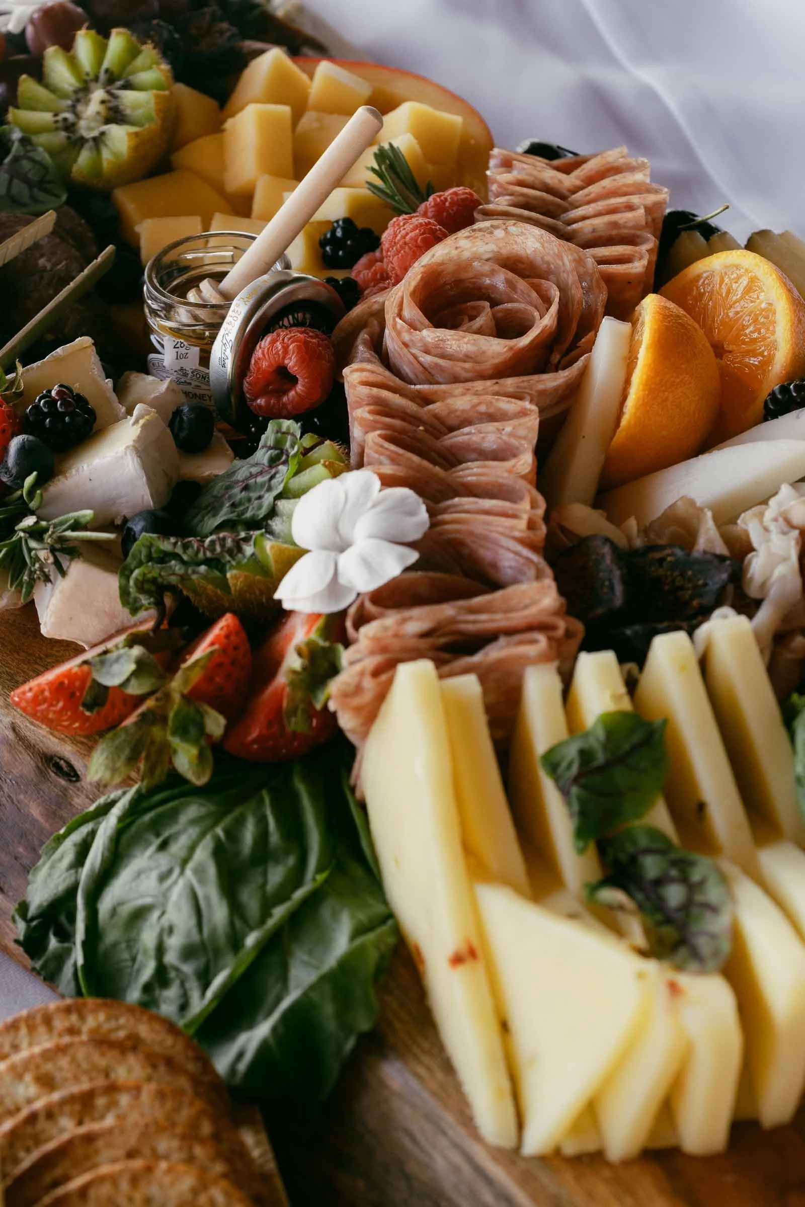  A wooden charcuterie board topped with sliced cheeses, cured meats arranged in a rose shape, fresh fruits including strawberries, kiwi, and oranges, leafy greens, crackers, and a jar of honey with a dipper. 