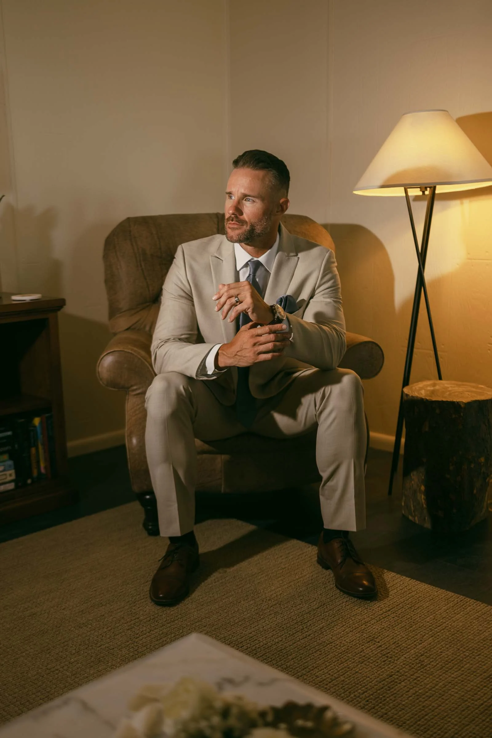  A man in a light beige suit sits on a brown armchair in a warmly lit room, looking to his left. He has short hair and a beard, and is holding his hands together. A floor lamp and a small bookshelf are in the background. 