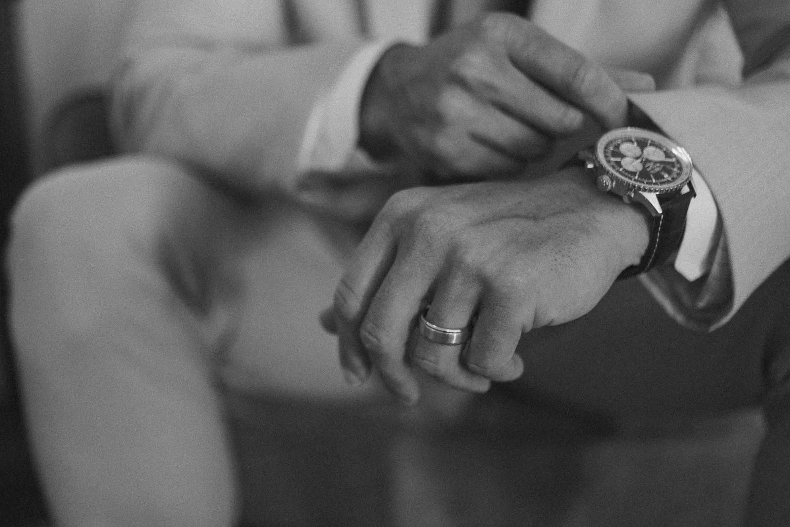  A person in a suit adjusts their wristwatch with one hand, while wearing a wedding ring on the other hand. The image is in black and white and slightly out of focus. 