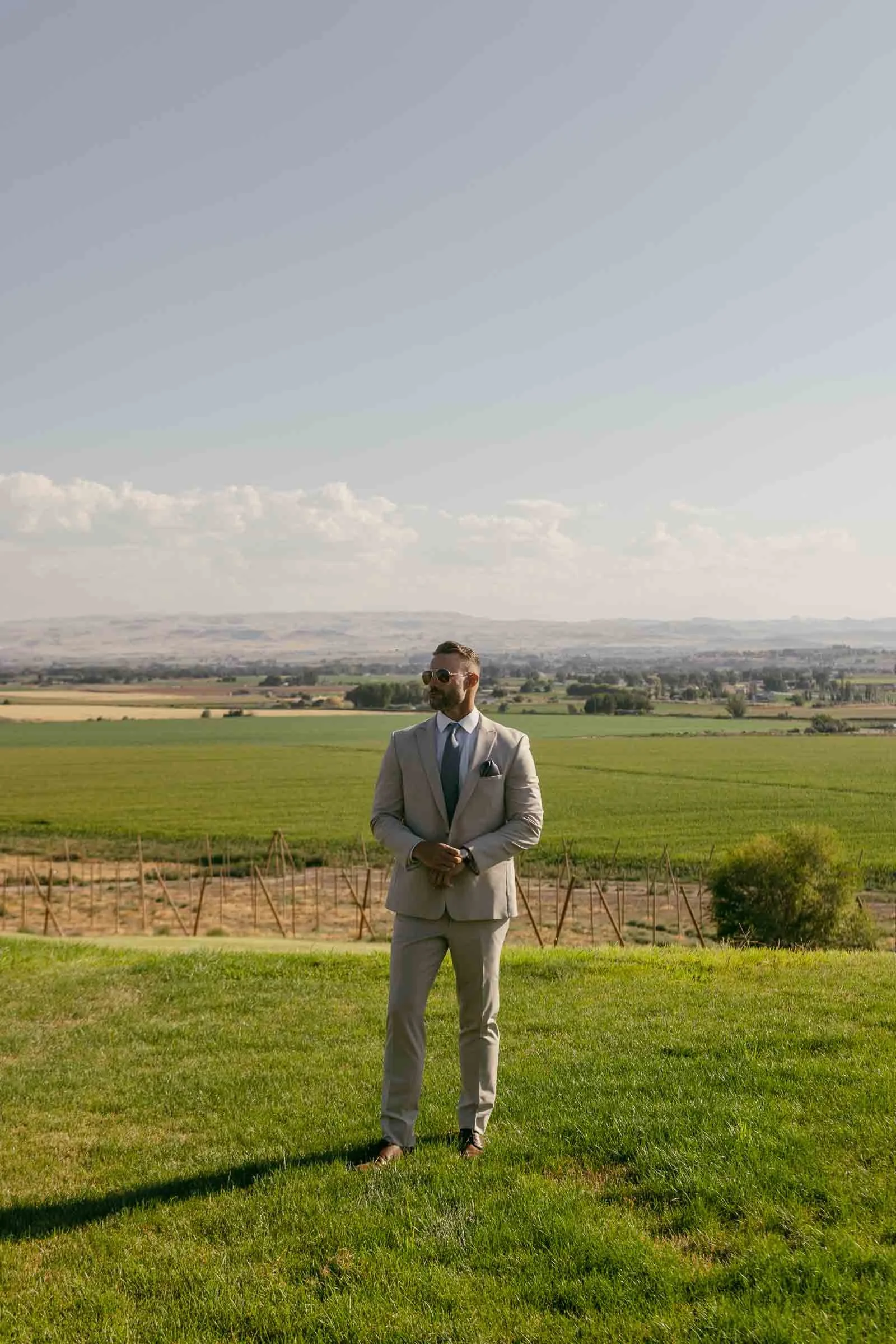  A man in a light-colored suit and sunglasses stands on a grassy lawn with fields and distant mountains under a clear sky in the background. 