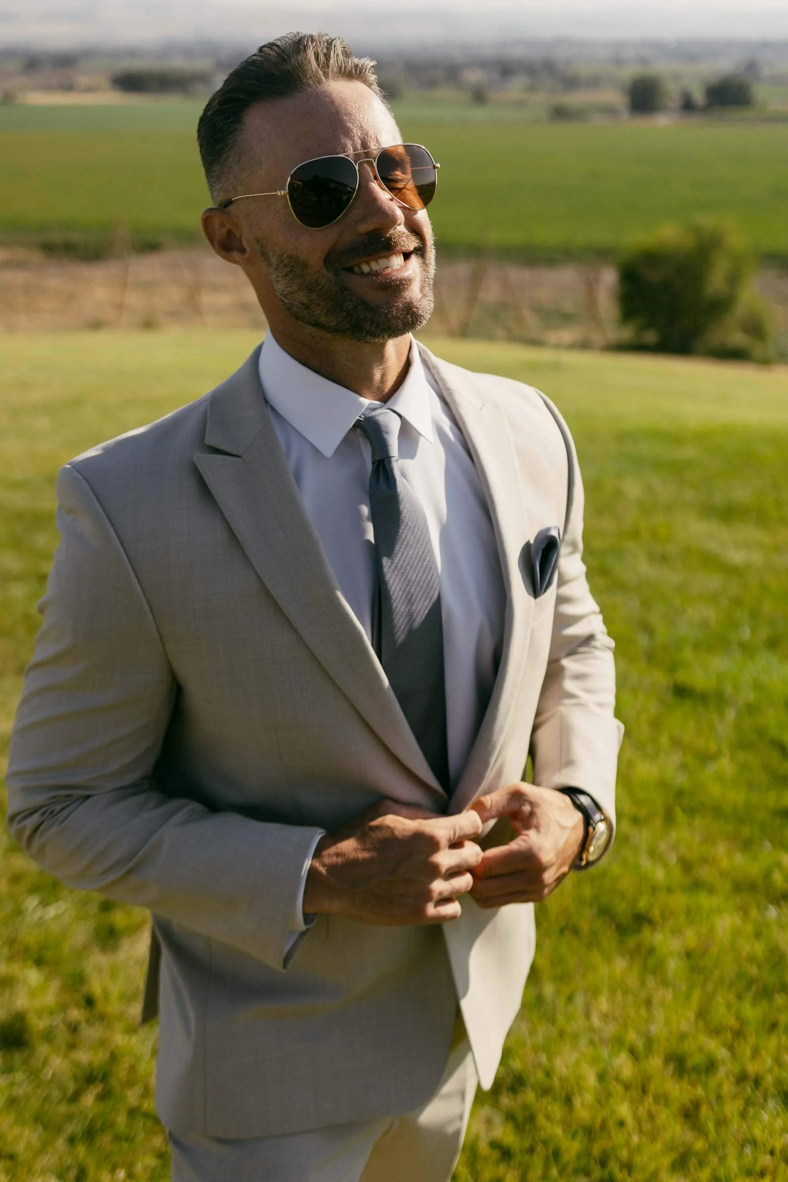  A man wearing a light beige suit, white shirt, and gray tie stands outdoors on green grass, smiling and wearing sunglasses. The background shows a scenic landscape with fields and distant trees. 