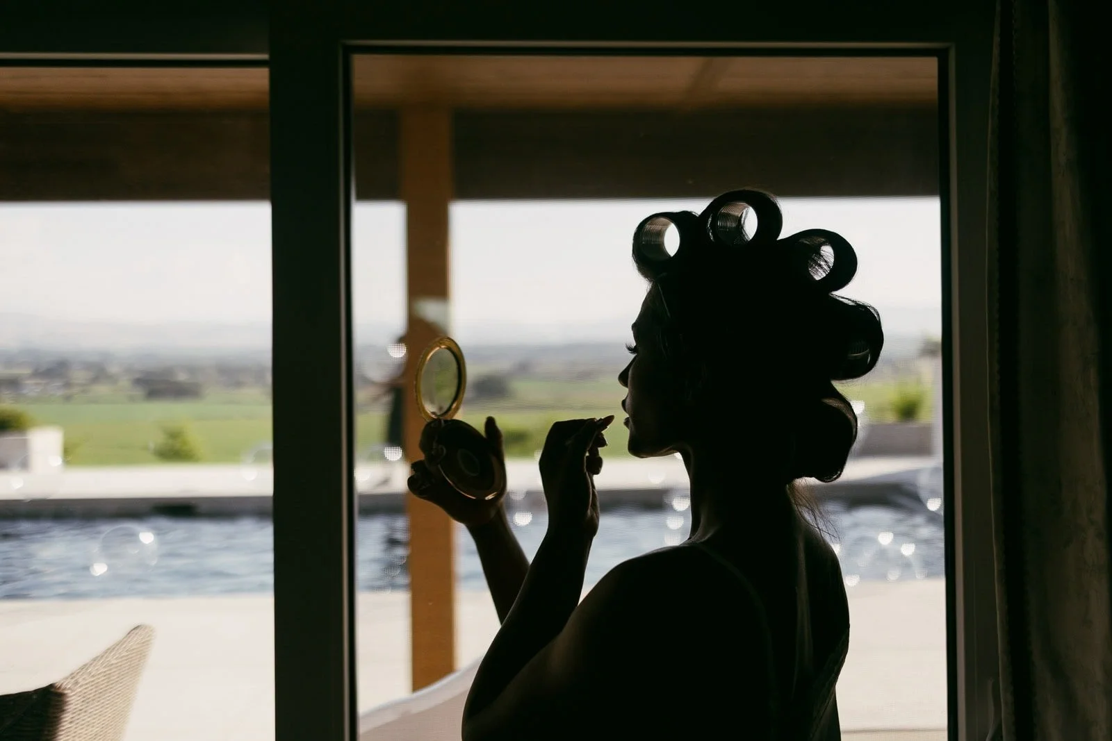  Silhouette of a person with hair rollers holding a compact mirror and applying makeup, standing near a window with an outdoor pool and landscape visible in the background. 