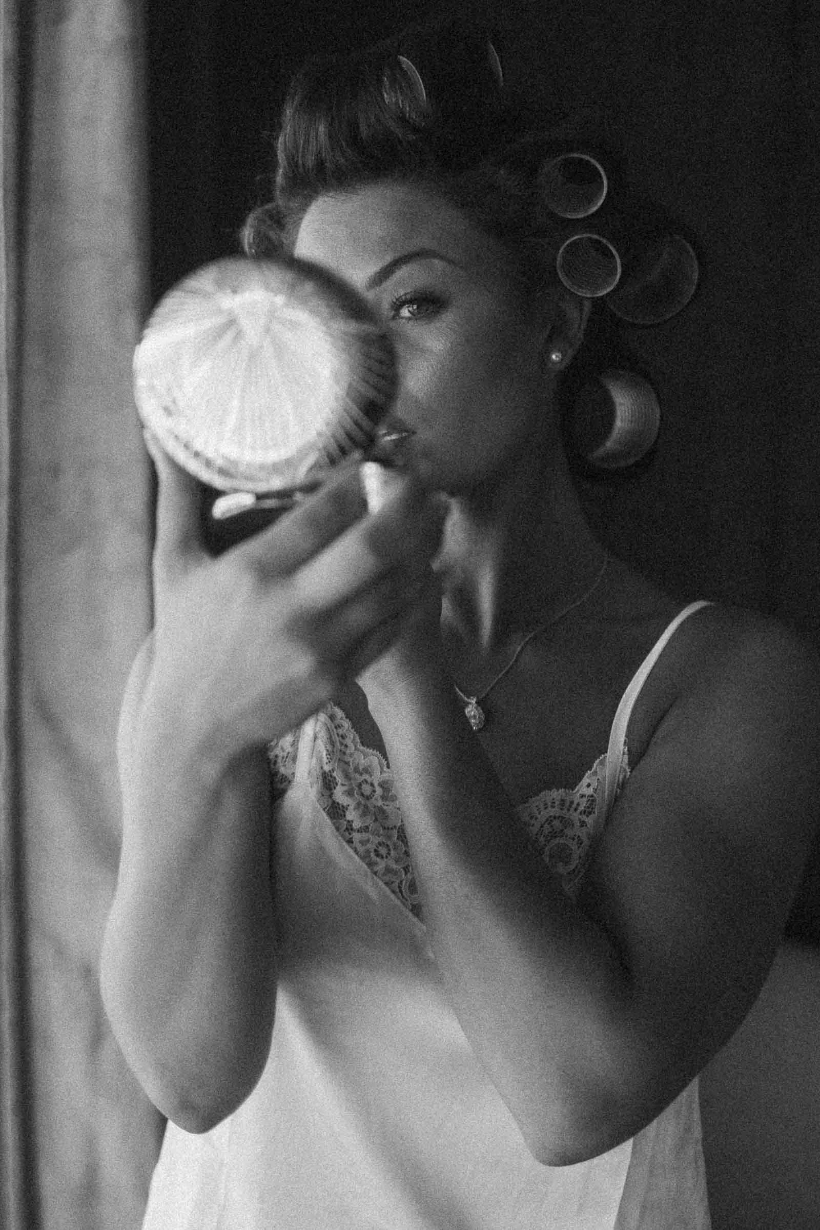  A woman in a lace-trimmed camisole holds a round mirror and applies makeup. She has hair rollers in her hair and looks intently at her reflection. The photo is in black and white. 