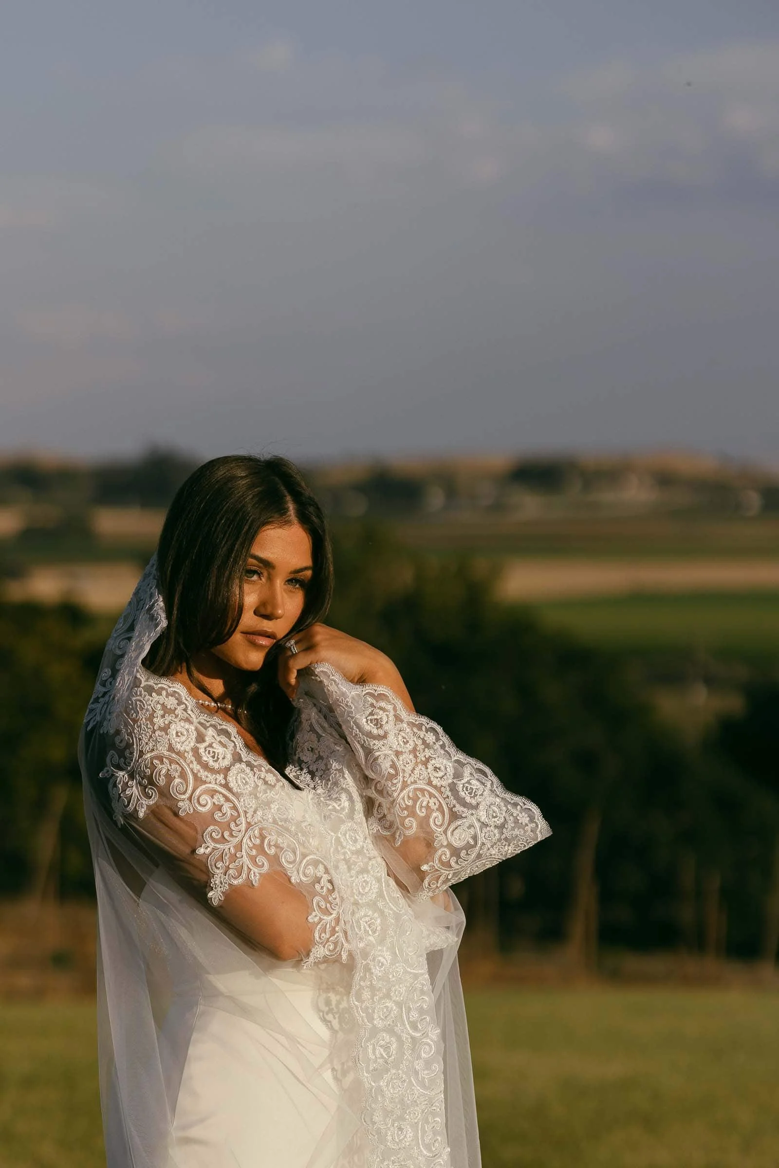  A bride in a white lace wedding dress and veil stands outdoors in a sunlit field with a soft background of trees and distant hills. 