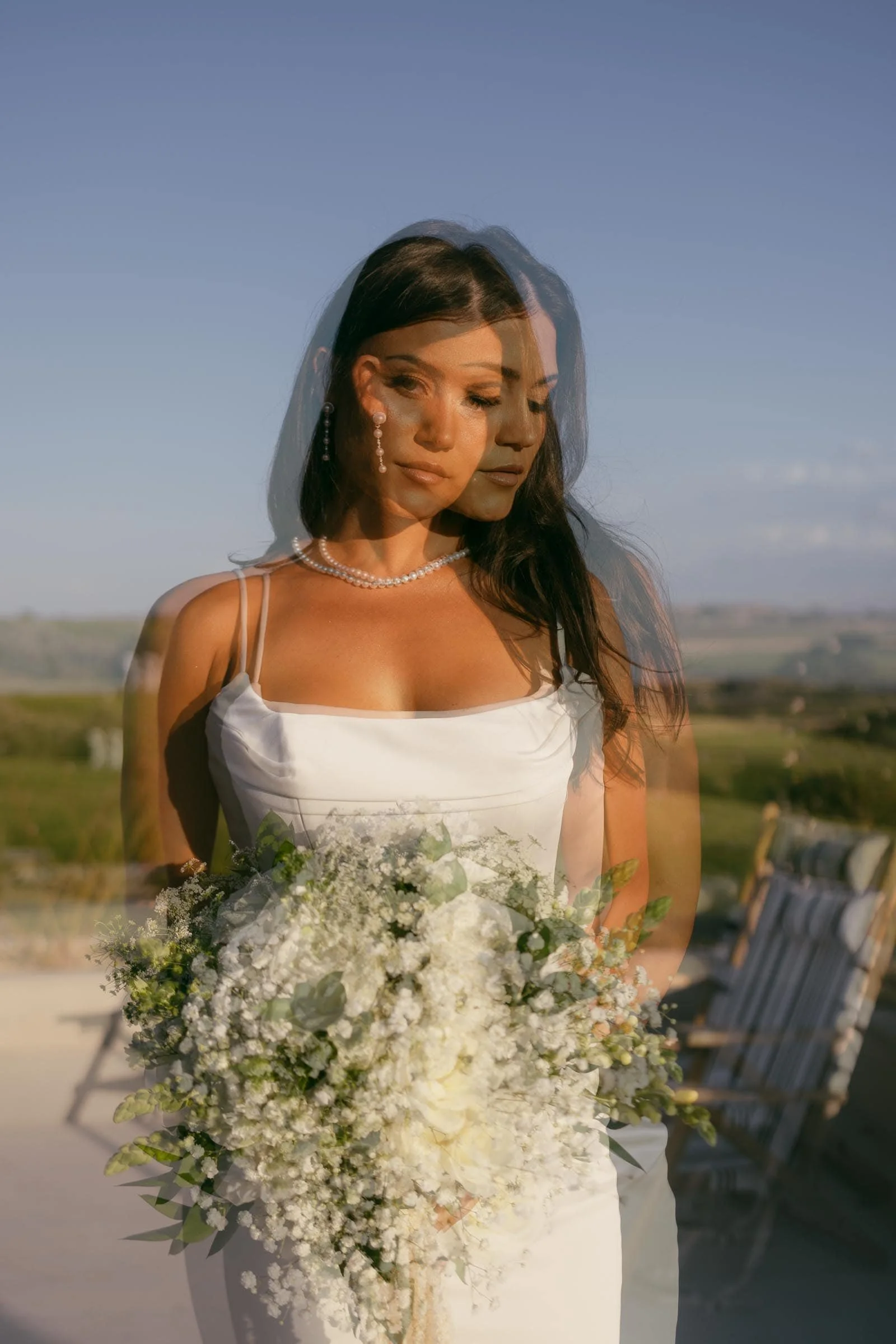  A woman in a white dress holding a bouquet of white flowers stands outdoors. The image has a double exposure effect, creating a ghostly, layered appearance. The background shows a scenic landscape under a clear sky. 