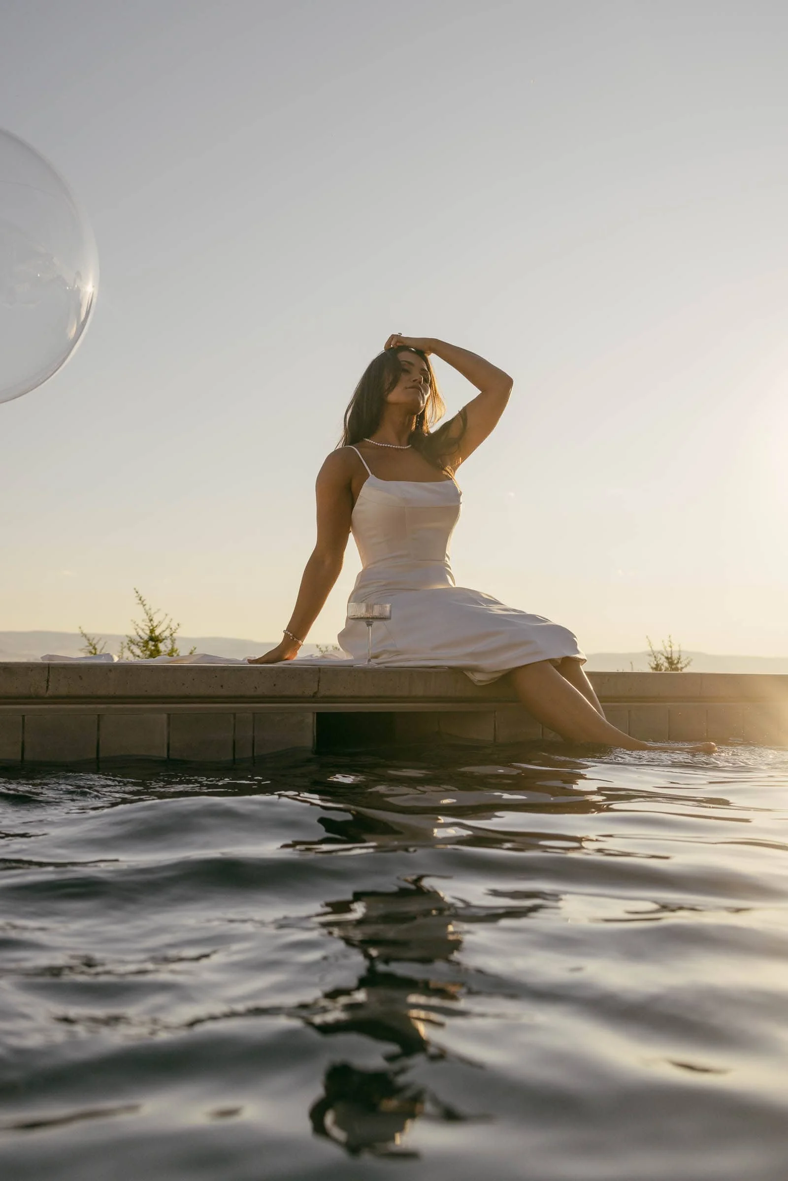  A woman in a white dress sits on the edge of a pool at sunset, with her feet in the water and one hand resting on her head. The scene is calm, with soft sunlight and gentle waves in the pool. 