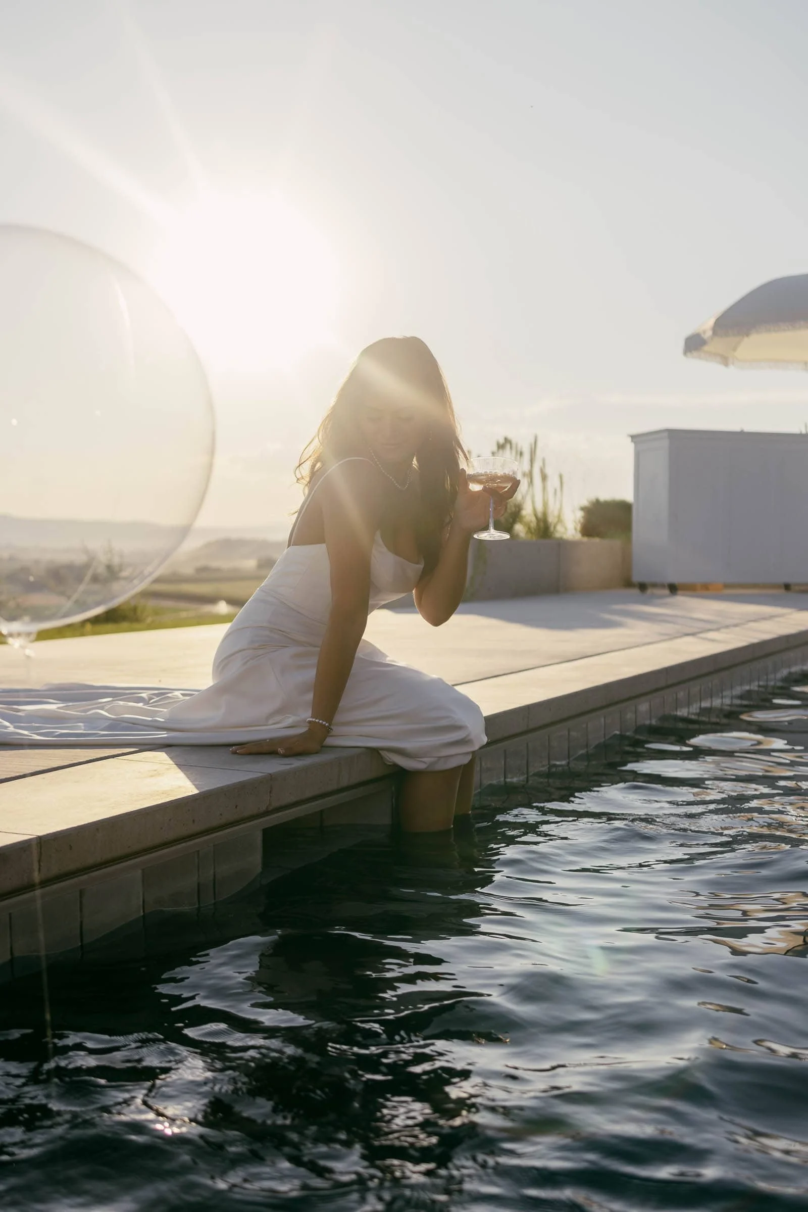  A woman in a white dress sits on the edge of a pool with her feet in the water, holding a drink and enjoying the sunset. The sun glows behind her, creating a warm, relaxed atmosphere. 