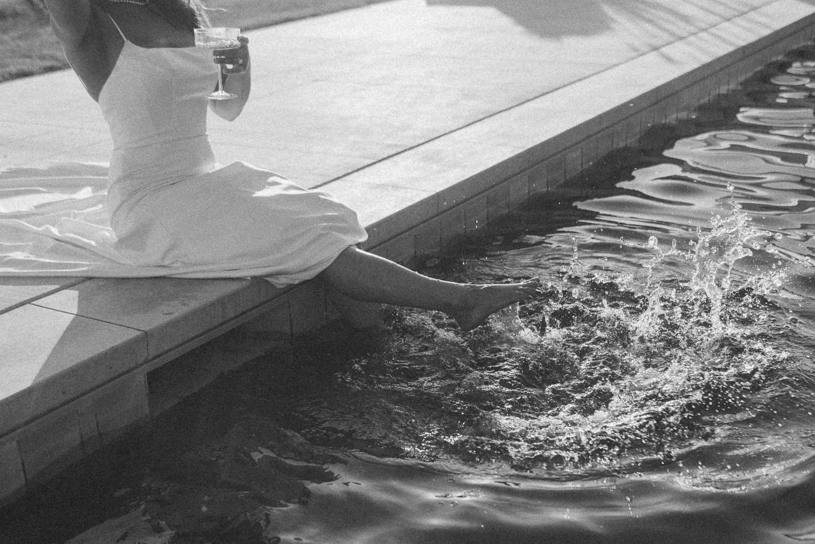  A woman in a white dress sits on the edge of a pool, holding a drink and splashing her feet in the water, creating ripples and splashes; the photo is in black and white. 