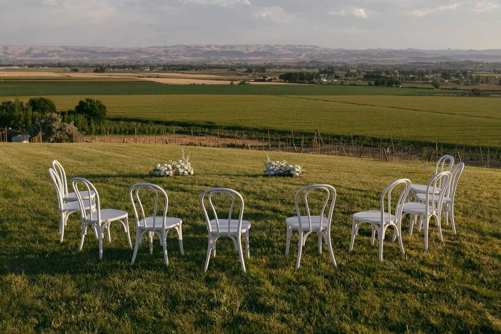  A small outdoor wedding setup with eight white chairs arranged in a semi-circle on a grassy hill, facing two floral arrangements, overlooking fields and distant hills under a partly cloudy sky. 