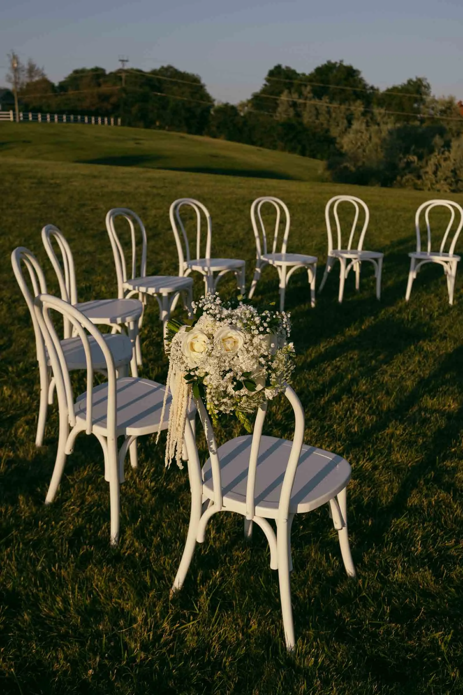  A group of white chairs arranged in a circle on a grassy lawn, with one chair decorated with a bouquet of white flowers and greenery. Trees and a fence are visible in the background under early evening light. 