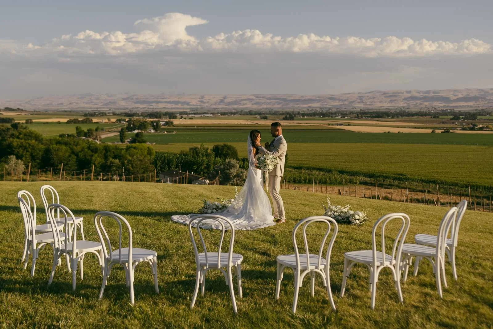  A bride and groom stand together on a grassy hill overlooking farmland and mountains, surrounded by a few white chairs arranged in a semi-circle for an intimate outdoor wedding ceremony. 
