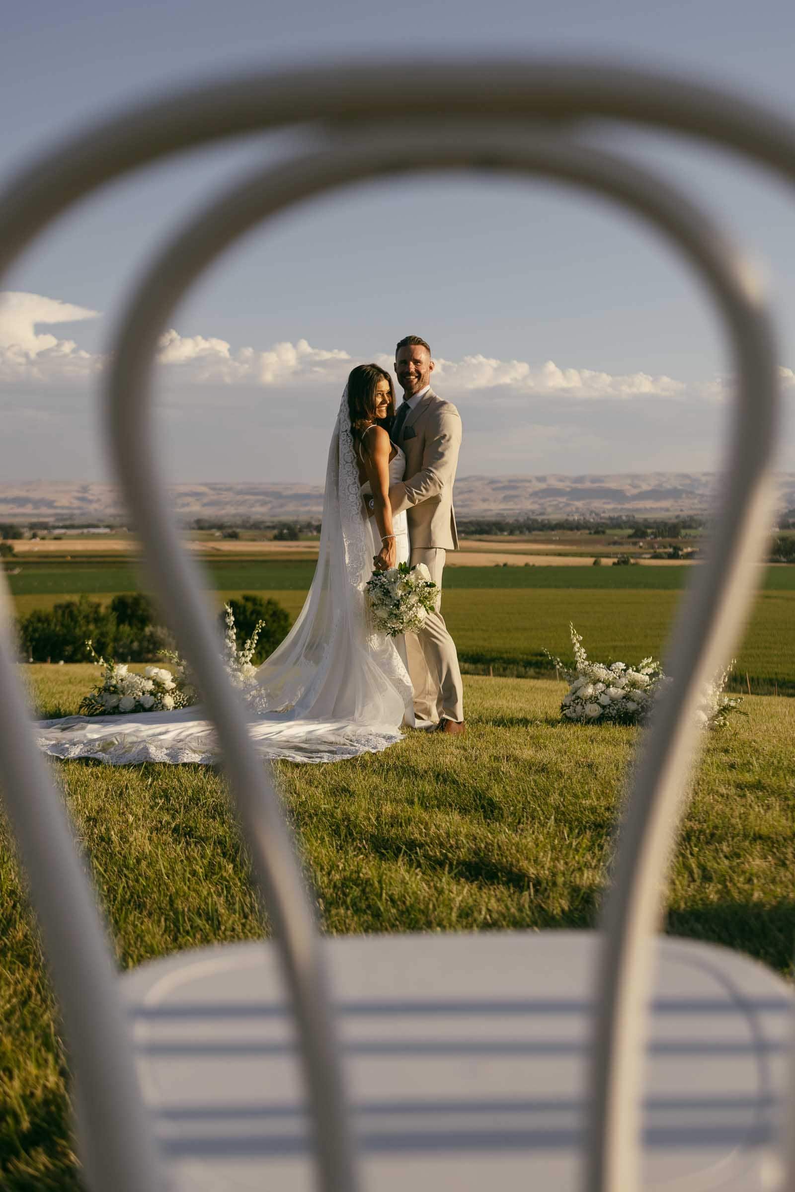 A bride and groom stand smiling on a grassy hilltop, holding a bouquet, with scenic fields in the background, framed creatively through the back of a white chair in the foreground. 