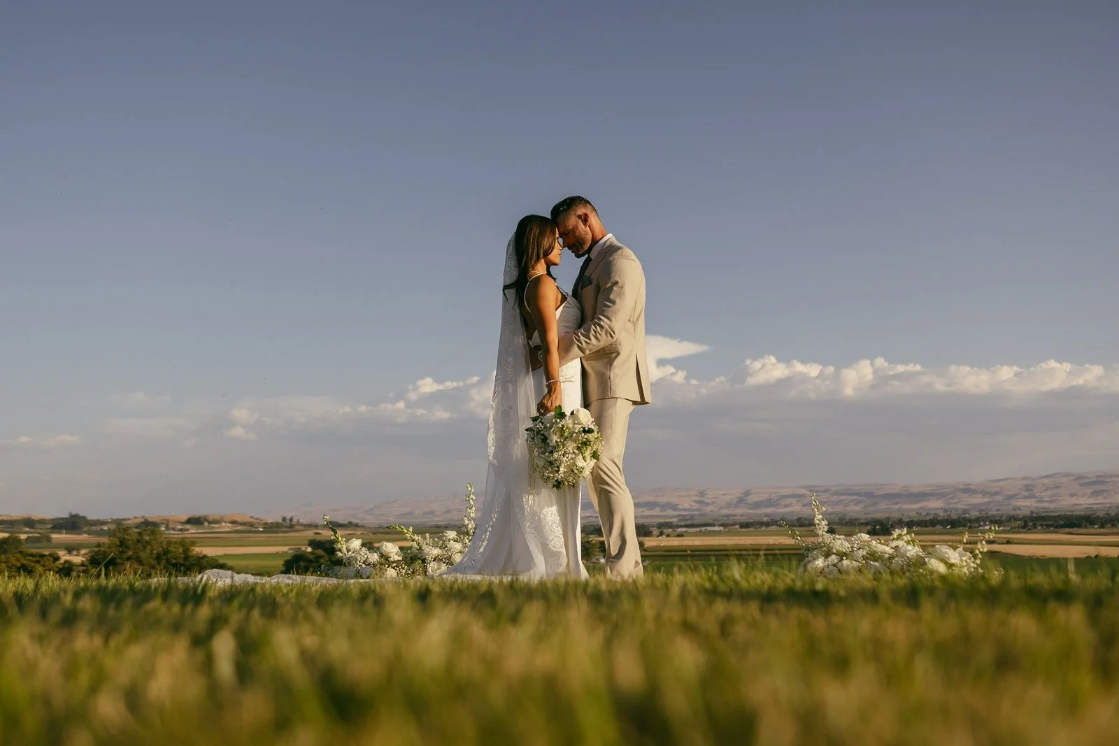 A bride and groom stand close together outdoors, touching foreheads and holding hands, with a bouquet of flowers. The background shows a wide landscape under a clear blue sky.