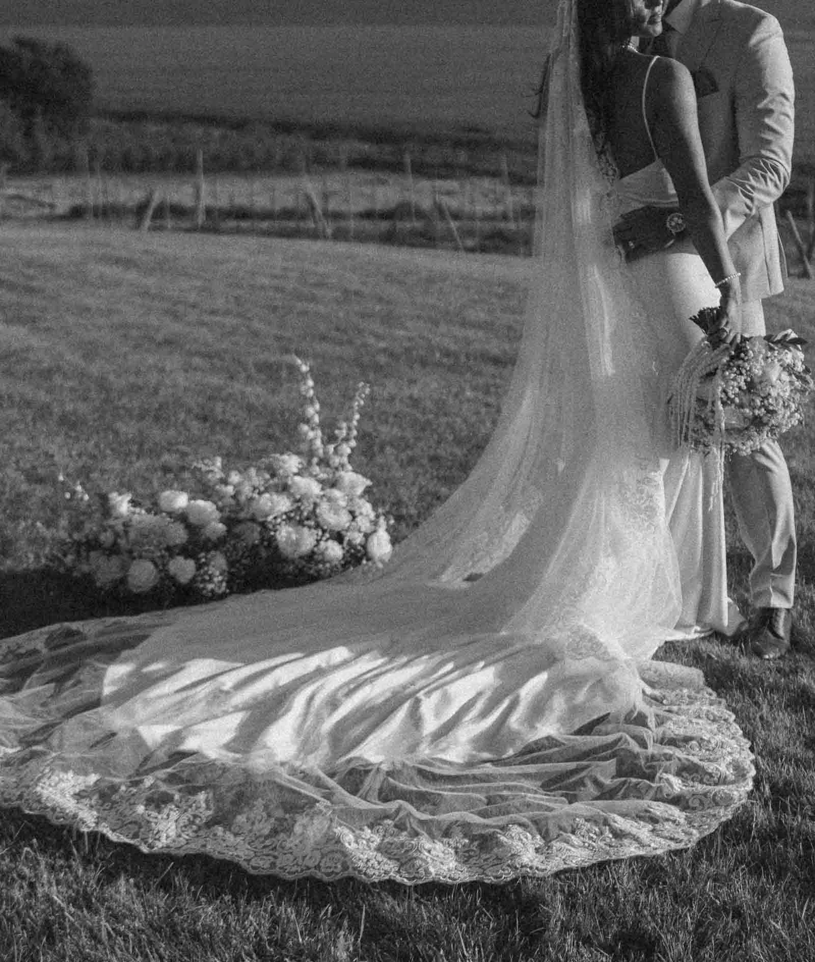  A bride in a long, lace-trimmed wedding dress and veil stands on grass beside a groom in a suit, holding a bouquet. A floral arrangement lies nearby, with open fields in the background. The image is in black and white. 